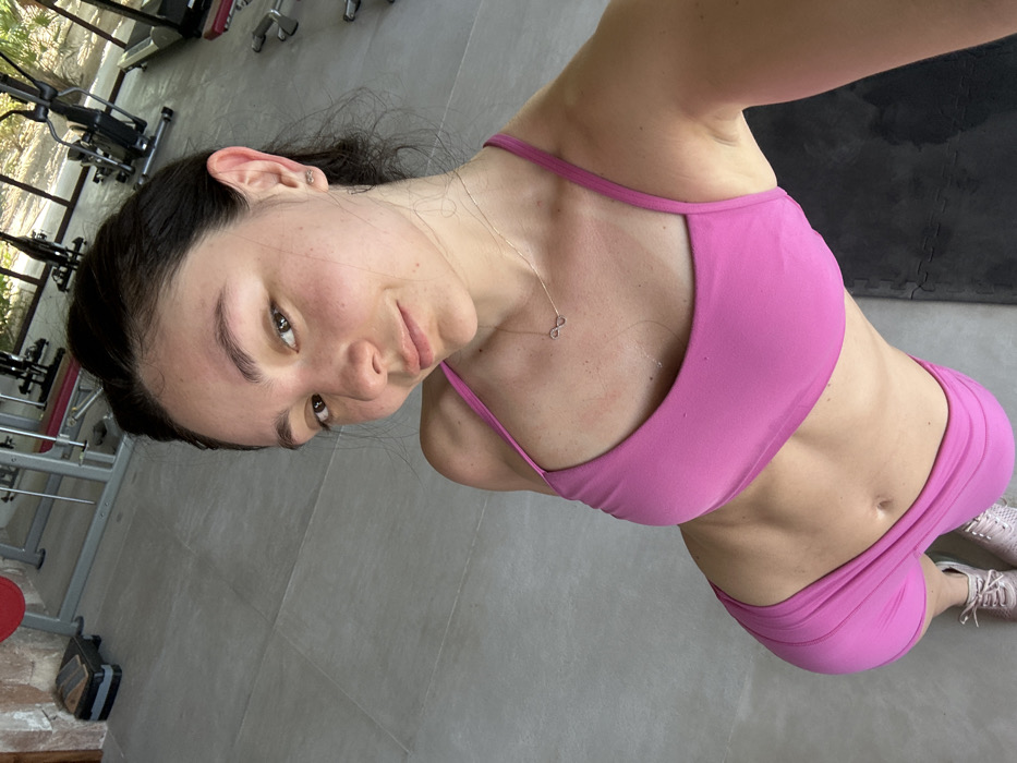 The image shows an adult female positioned in an indoor gymnasium located in Playa Blanca, Mexico. She is captured from a high-angle perspective, appearing to take a selfie. She has dark hair and fair skin with visible freckles, and is wearing a pink sports bra, matching pink athletic shorts, and light-colored athletic shoes. A silver necklace with an infinity symbol pendant and small infinity symbol stud earrings are visible. Her midsection displays muscle definition. The background reveals various gym equipment, including a weight rack with red weights, multiple treadmills, exercise bikes, and a black stepper. The floor consists of large, light grey square tiles, with a black mat visible under her feet on the right. A stone wall section is present on the far left, and foliage from outside can be seen through open or windowed sections in the background, suggesting natural light.