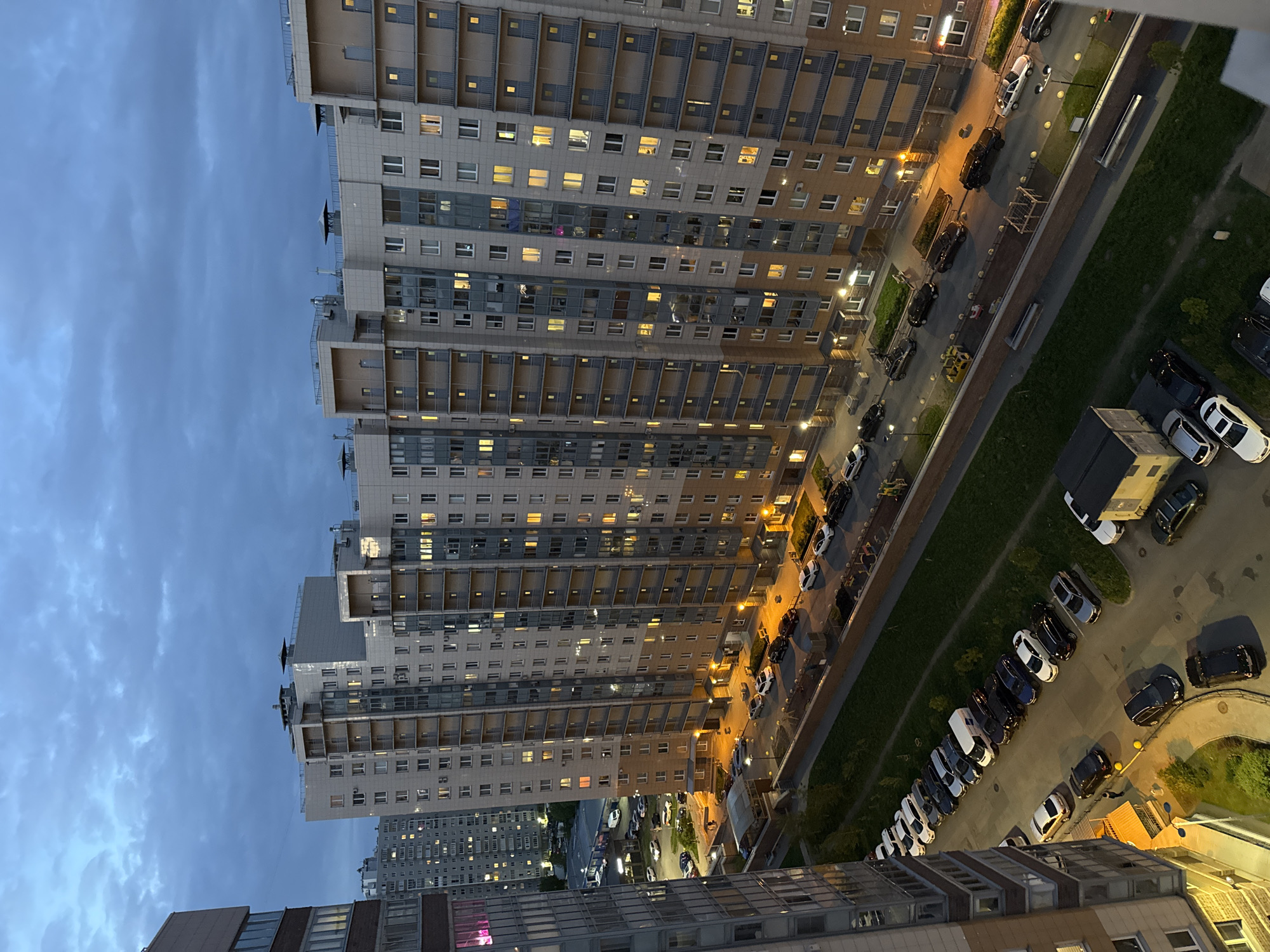 The image is a high-angle, nighttime shot of a dense urban environment, seemingly from the top of a building. The buildings, a mixture of gray and light beige, are modern high-rises with multiple floors and numerous windows, many of which are illuminated. A street below is lined with parked cars and sparse greenery, suggesting a residential area. Streetlights cast a warm, orange glow, illuminating the parked cars and the surrounding pavement. The sky above is a dusk blue, hinting at the time of day. The overall atmosphere is calm and urban, with a sense of residential tranquility despite the density. No people are readily visible in the photograph.