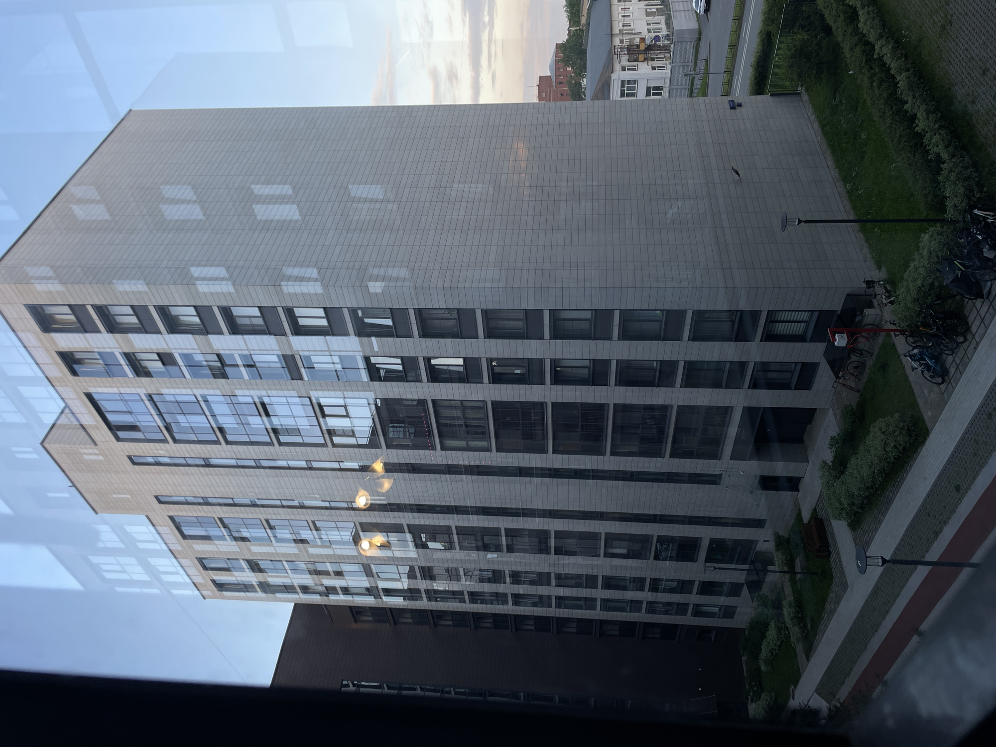 The image shows a modern apartment building in Sinyavino, Russia, viewed from a window. The building is tall and rectangular, with a facade of light-colored tiles and numerous windows. There are reflections on the window glass, including the sky and other buildings. In the foreground, there is a courtyard area with paved walkways, grassy patches, and bushes. Several bicycles are parked near the building, and a bench is visible in the courtyard. Beyond the apartment building, other structures and trees can be seen, with a road and a white car in the distance. The sky appears partly cloudy, with hints of sunset colors. The overall scene depicts a residential urban environment.