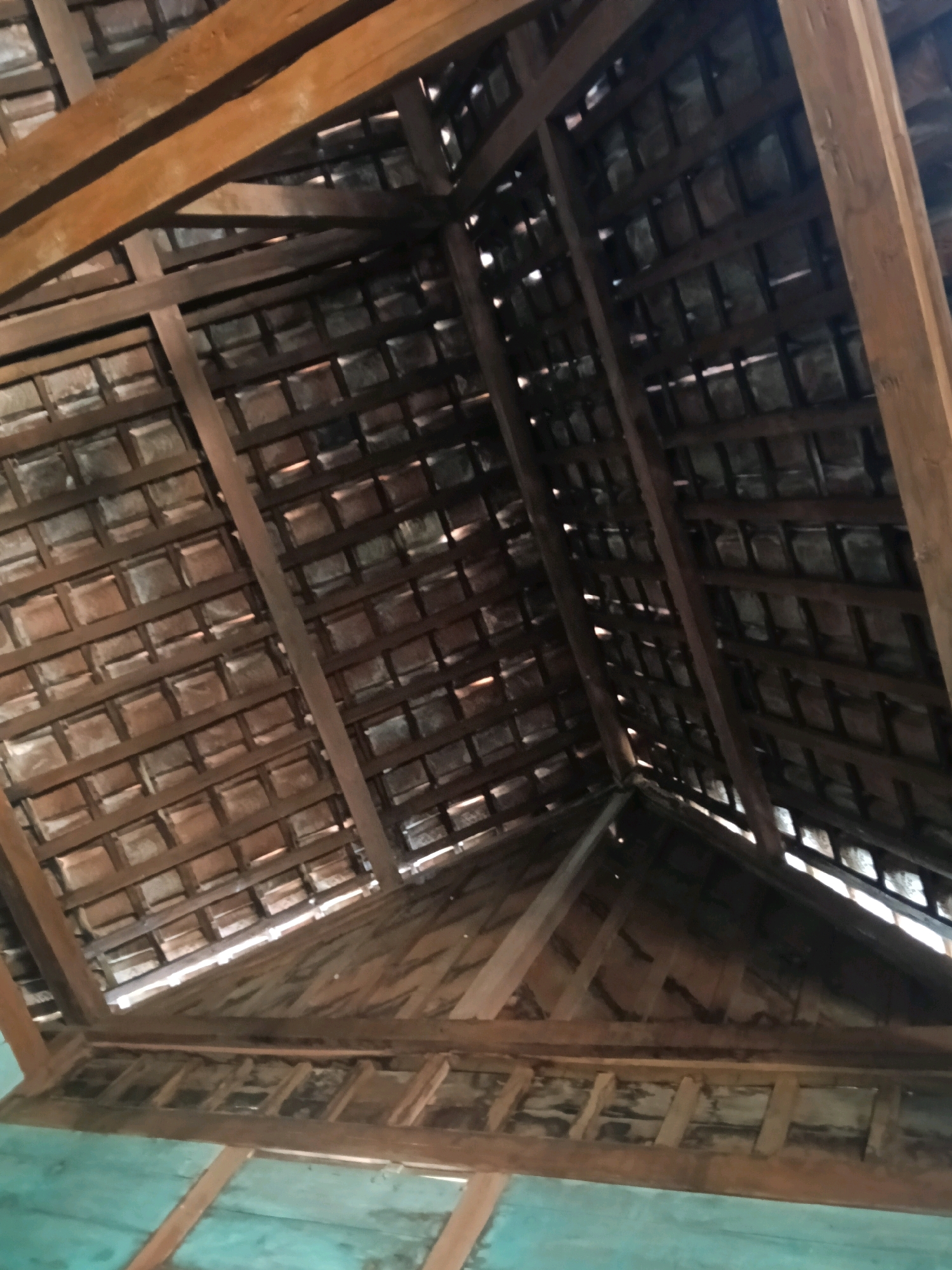 The image shows the underside of a wooden roof structure. The roof is constructed with visible wooden beams and rafters, supporting rows of dark, rectangular roof tiles. Gaps between the tiles allow light to filter through, creating a patterned illumination. In the foreground, a turquoise-painted wooden wall or ceiling is partially visible. The scene appears to be an interior view of a building, likely a traditional dwelling given the materials. The location is identified as Nglungger, Indonesia. There are no people or animals present in the image.