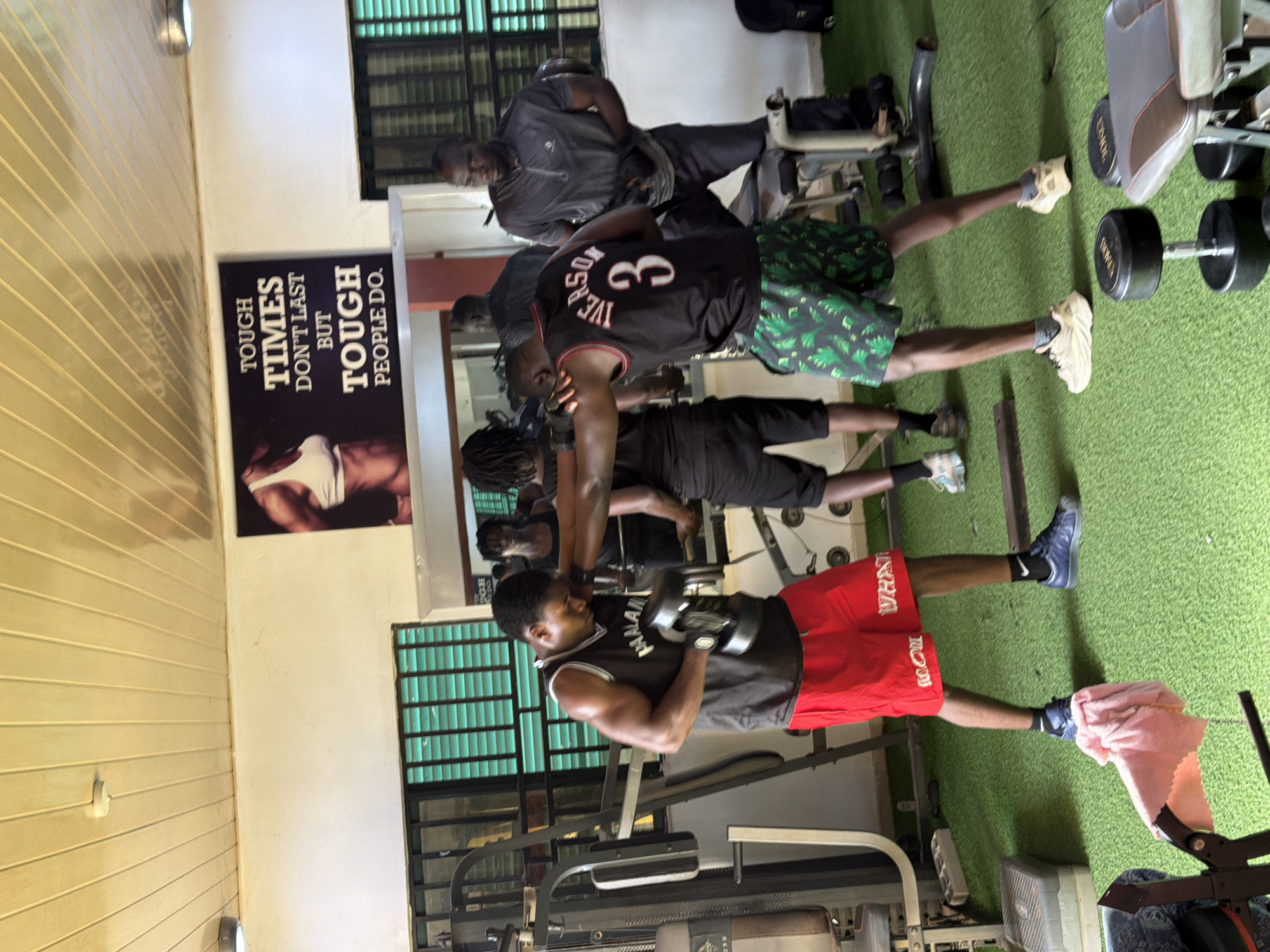 The image depicts the interior of a gym located in Gura Top, Nigeria. Two men are engaged in a boxing-like training session in the foreground, with one man wearing red shorts and a black tank top, holding a dumbbell, and the other wearing patterned shorts and a black tank top with the name "IVERSON" and the number "3" on the back. Several other individuals are visible in the background, some appearing to be observing or waiting. The gym is equipped with various weight machines and dumbbells, with a bright green carpeted floor. A motivational poster with the text "TOUGH TIMES DON'T LAST BUT TOUGH PEOPLE DO" is displayed on the wall above a mirror. The overall environment suggests a functional, albeit somewhat rustic, fitness facility.