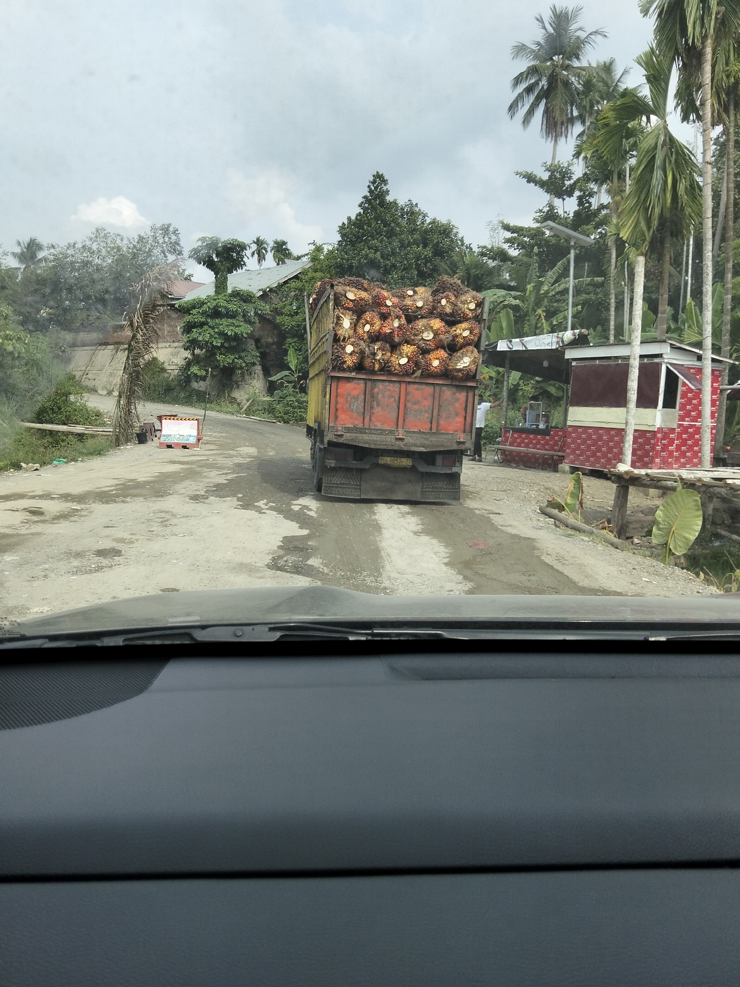 The image captures a scene on a rural road in Awegeutahpaya, Indonesia, likely a palm oil plantation area. A large, red and yellow truck is prominently featured, heavily laden with fresh palm oil fruit bunches. The truck is on a slightly wet, unpaved road that winds through a lush, green environment with numerous palm trees and dense foliage. To the right of the road, a small, open-air structure with a red and white patterned facade is visible, possibly a roadside stall or kiosk. A person in a white shirt is standing near this structure. In the background, there are several buildings with roofs, suggesting a small settlement or part of the plantation infrastructure. The sky is overcast with clouds. The perspective suggests the photo was taken from inside a vehicle, looking forward through the windshield.