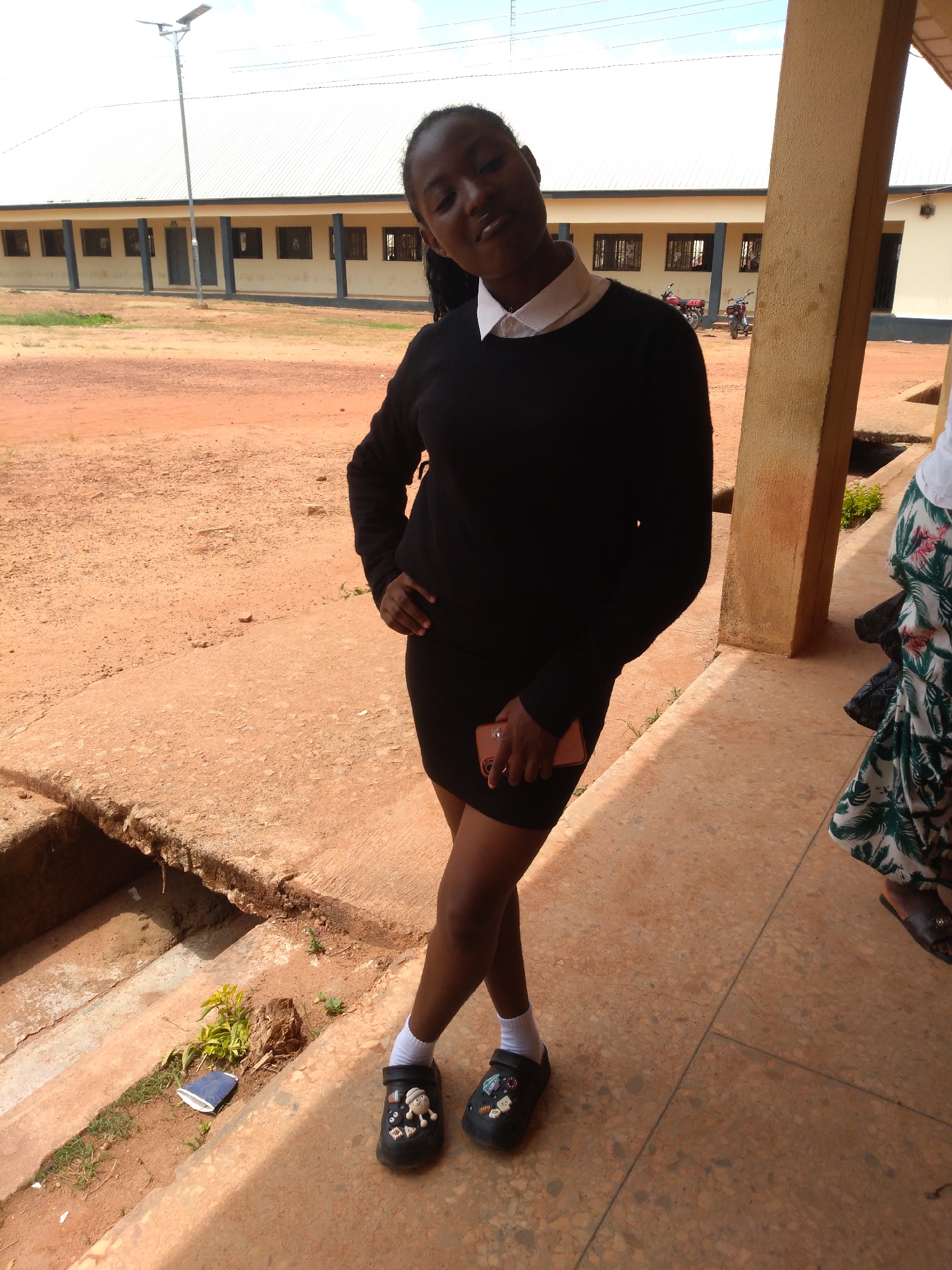 A young woman stands with her left hand on her hip and her right hand holding a pink smartphone. She is wearing a black sweater over a white collared shirt, a black skirt, white socks, and black clogs adorned with cartoon figures. The background features a light-colored building with windows and a covered walkway, suggestive of a school or administrative facility. To the right, another person's legs are visible, wearing a white skirt with a green and white tropical pattern and brown sandals. The ground is covered in reddish-brown dirt and concrete. In the distance, two motorcycles are parked. The scene appears to be outdoors, possibly in a courtyard or an area adjacent to a building. The lighting suggests it is daytime. This location could plausibly be in Jos, Nigeria, given the architectural style and general environment.