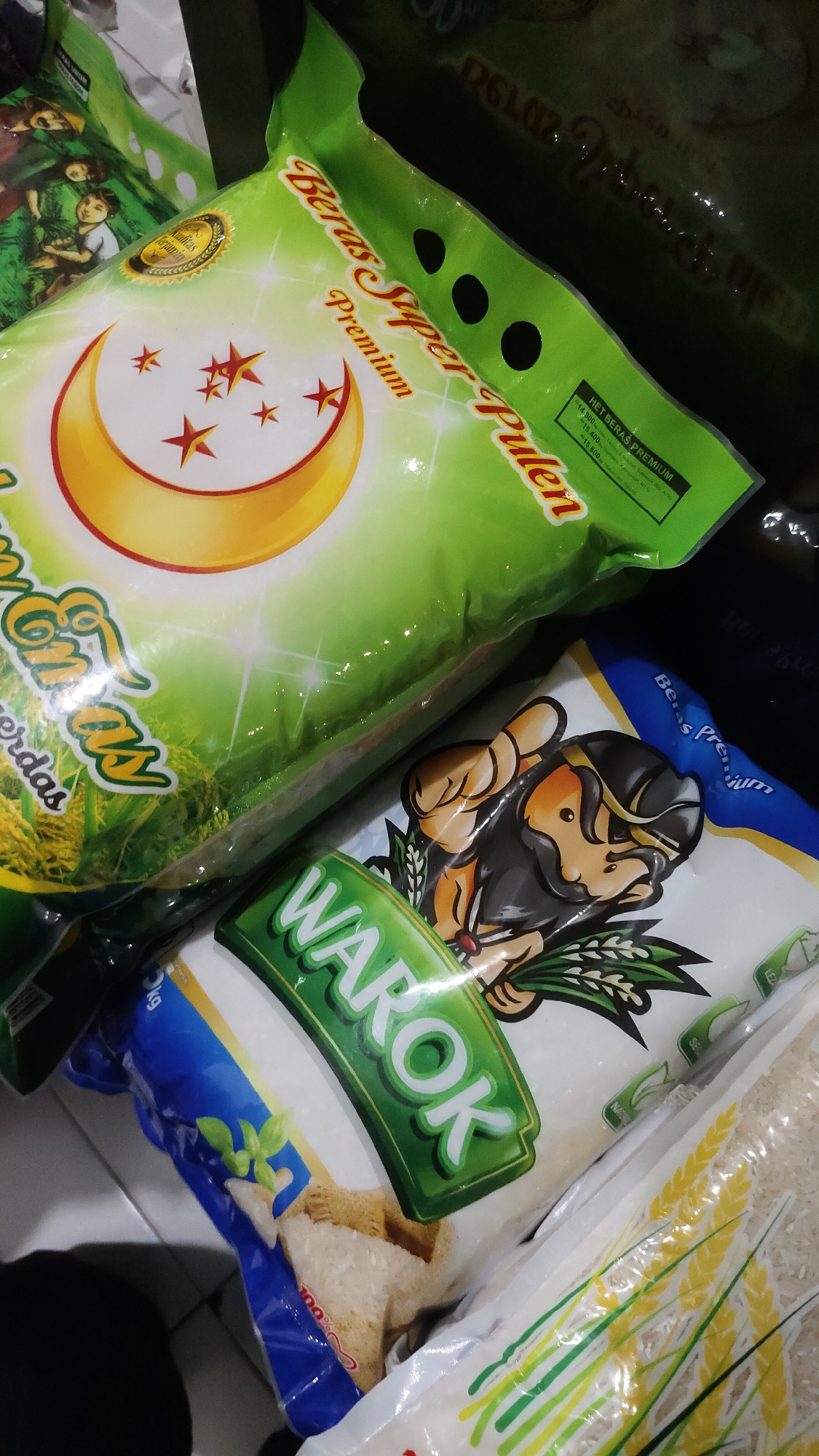 This is a close-up, high-angle, eye-level shot of several bags of rice arranged on a dark surface, likely a store shelf or floor, in Malang, Indonesia. The lighting is dim, and the surfaces are reflective, indicating a manufactured environment, possibly a retail setting.
Dominating the foreground is a large, green bag of rice with a prominent yellow crescent moon and red stars design. Text on this bag reads "Beras Super Pulen Premium." Another bag, partially obscured, features a cartoon illustration of a man with a traditional Javanese headdress. A prominent green label on this bag displays the word "WAROK" in large white letters. This bag is also white with blue accents. Other bags of rice, mostly out of focus and largely unidentifiable, are visible in the background. The overall impression is of a retail display of rice products.