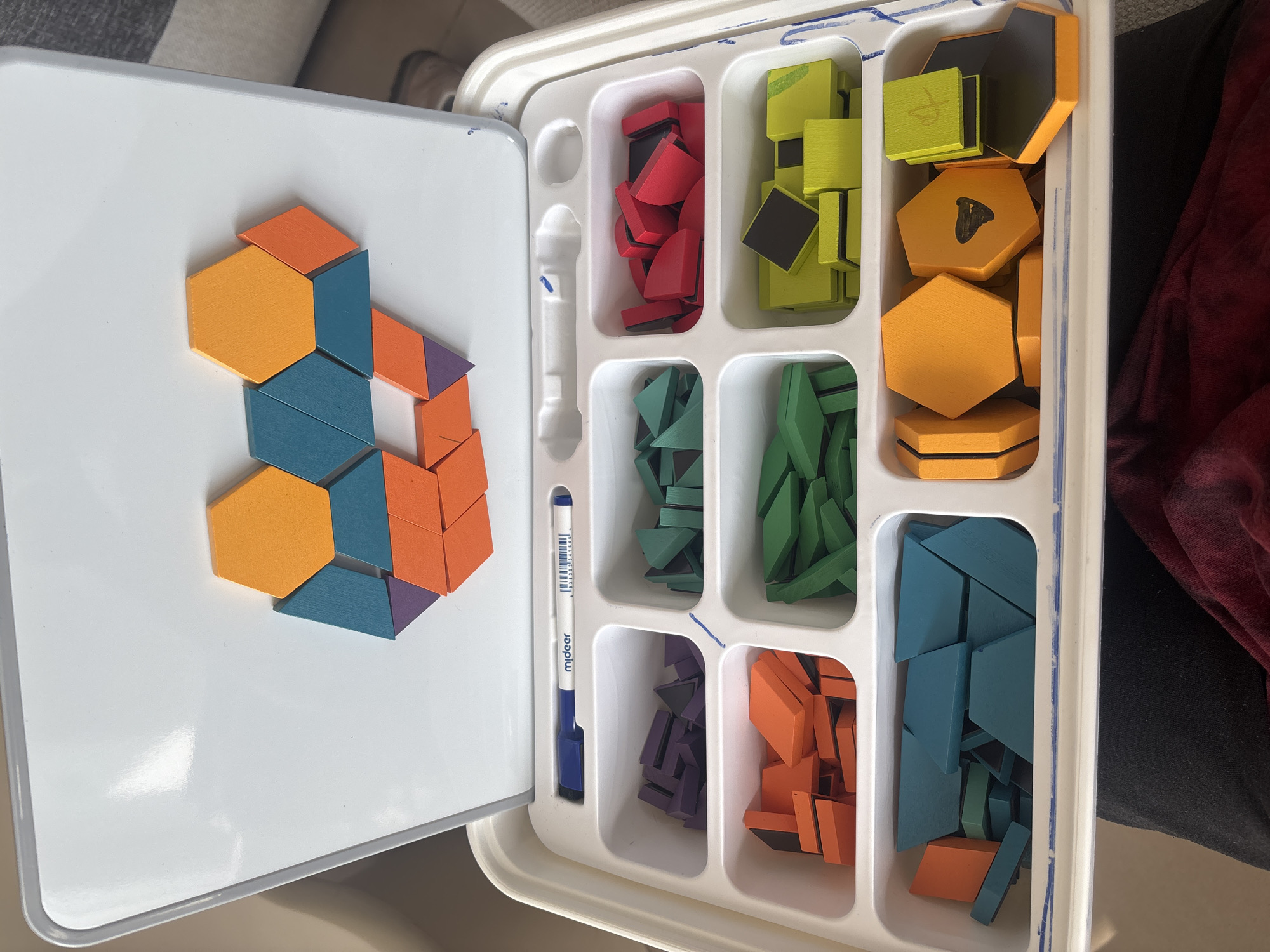 An overhead, medium shot shows a disassembled geometric shape-building toy set arranged in a white plastic tray with a white lid opened upwards. The lid displays a partially completed geometric pattern composed of orange, teal, and purple polygonal wooden pieces. The tray is divided into six compartments, each containing different colored sets of wooden geometric shapes. From the top row, left to right, the compartments hold purple, green, and red shapes. The bottom row, left to right, contains teal, yellow, and orange shapes. A blue marker with the brand "mideer" is placed in a designated slot above the compartments. The background is blurred and shows indistinct indoor surroundings. The image does not contain any identifiable landmarks or features that would indicate a specific location in Dubai, United Arab Emirates.
