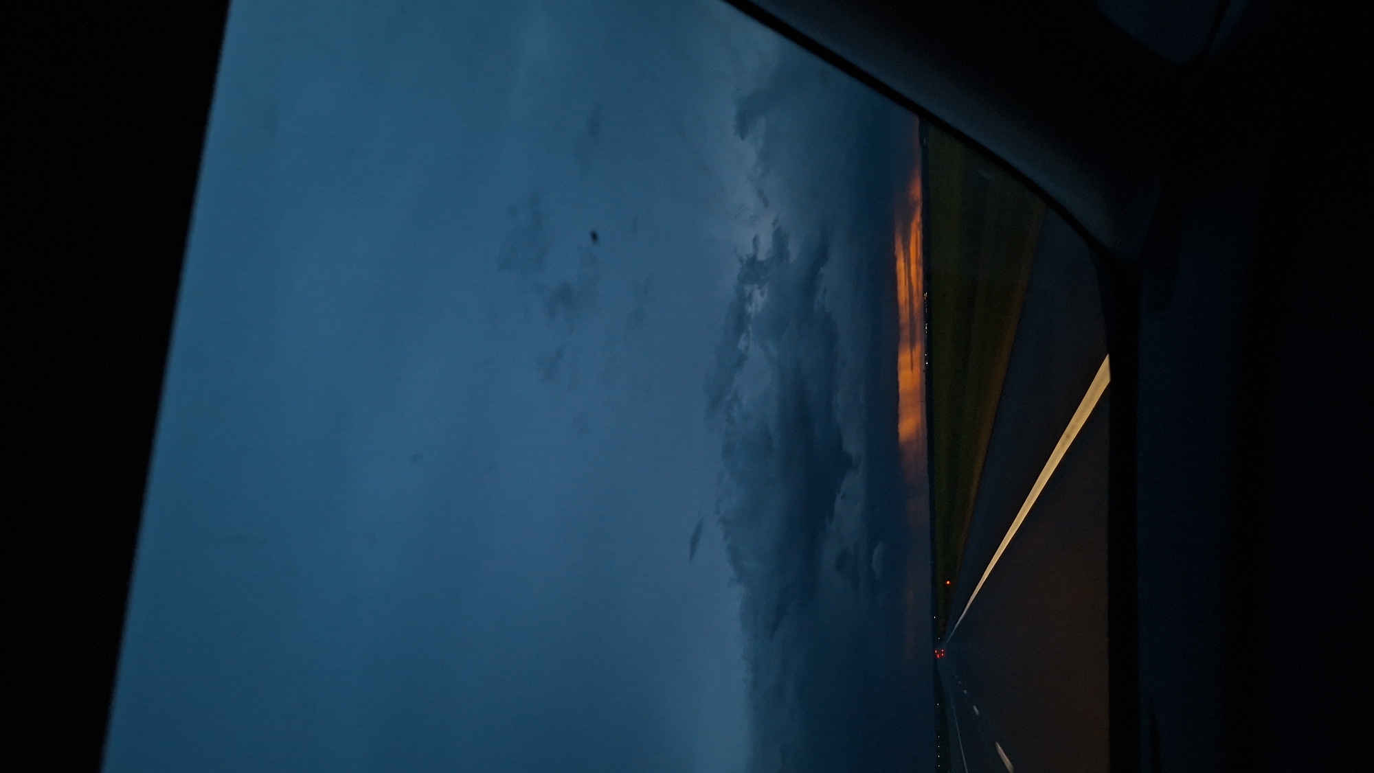 A view from inside a vehicle at night on a road in Дабрынёўскі сельскі Савет, Belarus. The sky is dark with heavy clouds, reflecting an orange hue on the horizon. A blurred highway with a painted white line is visible through the window.