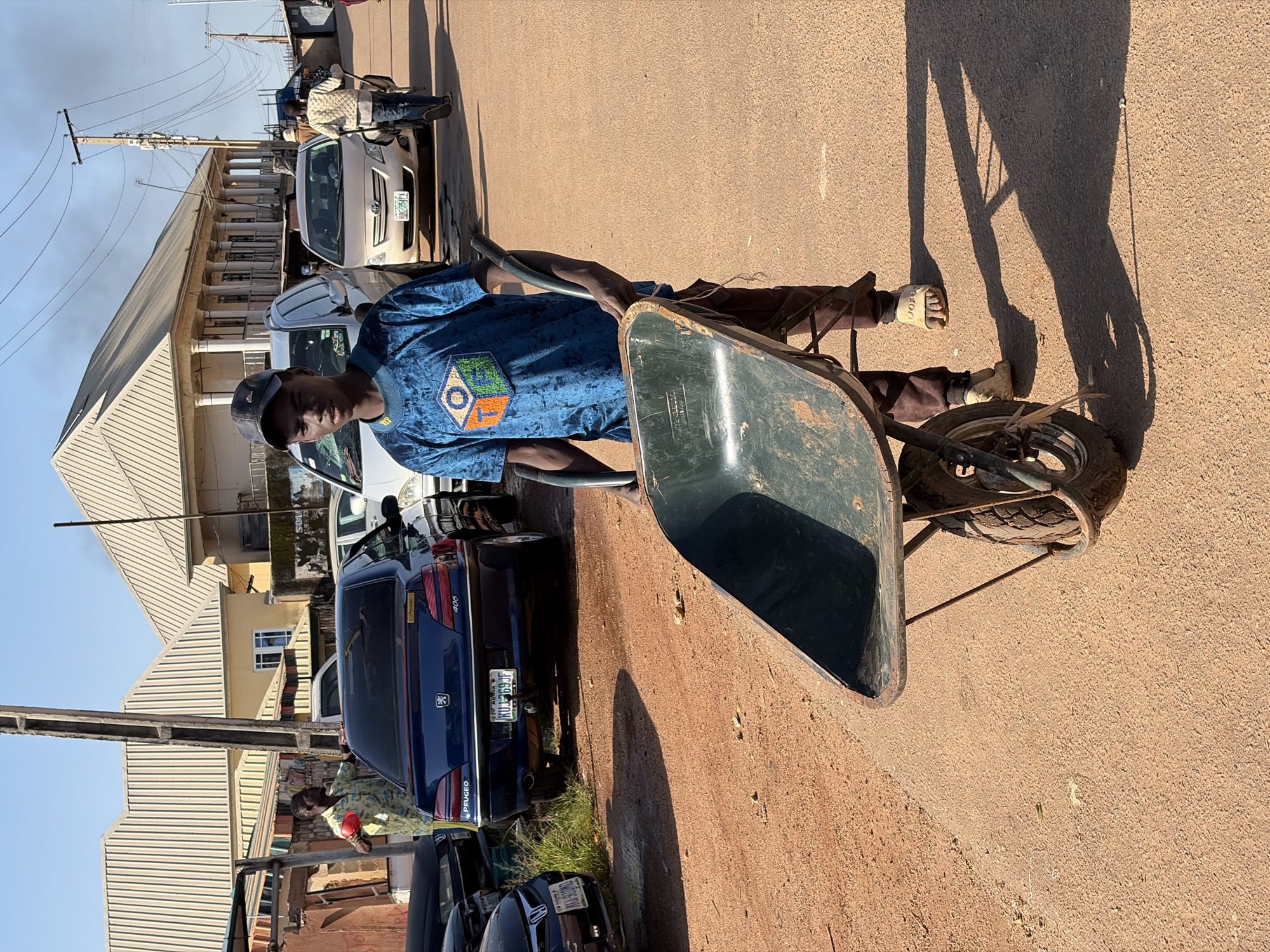 A young Black male, wearing a blue t-shirt with an emblem and brown pants, pushes a green wheelbarrow on a dusty, reddish-brown road. He is wearing sandals. The scene appears to be a street in Gura Top, Nigeria. In the background, several cars are parked or driving. A light-colored car is in the center background. To the left, a dark blue Peugeot 406 sedan is parked. Behind the young man, a person in a patterned jacket is walking away, also pushing a wheelbarrow. Buildings with corrugated metal roofs are visible on either side of the street. Power lines and utility poles are present overhead. The sky is clear and blue, and the sun casts long shadows on the ground.