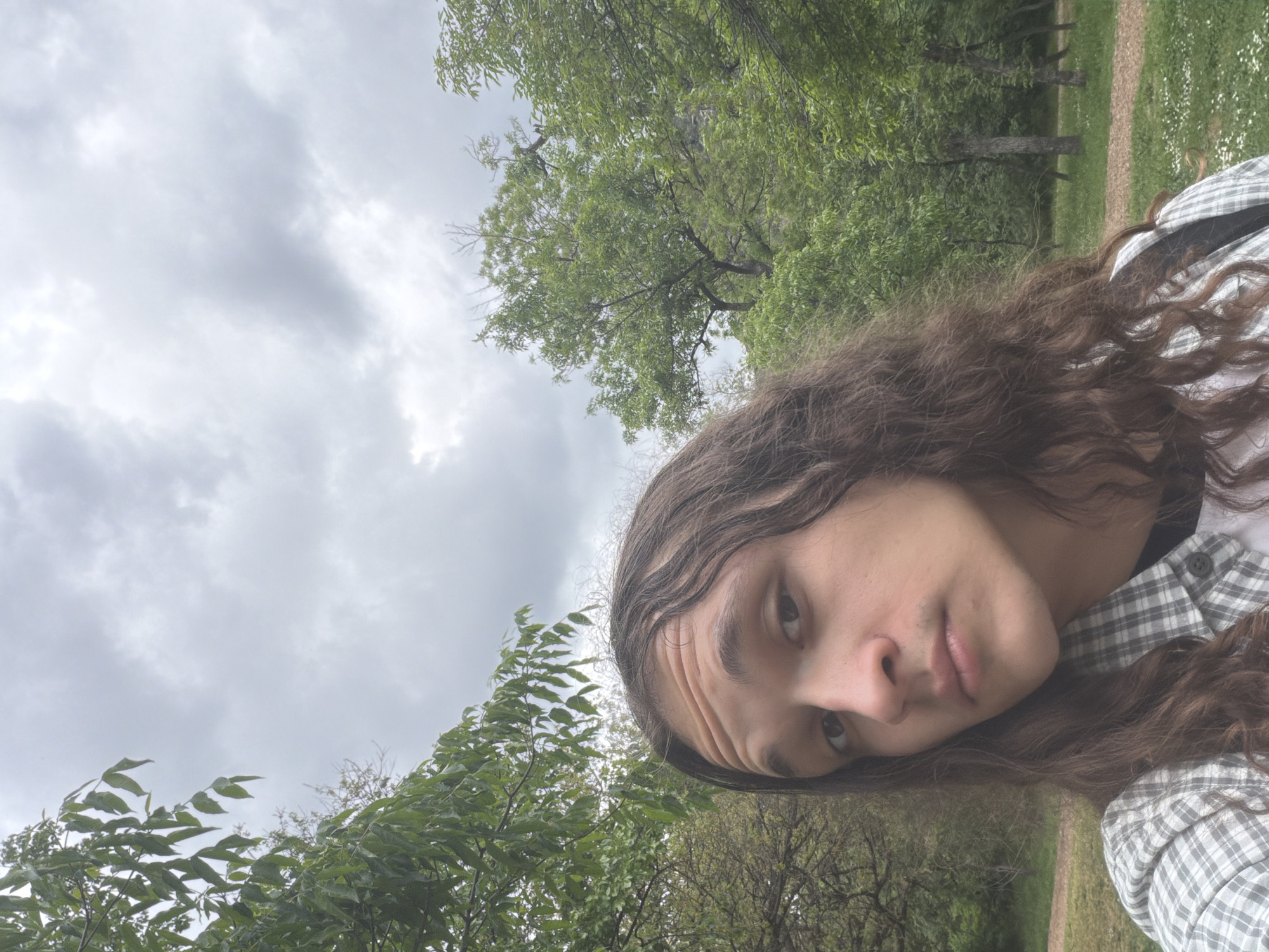 A sideways selfie taken in a park. The subject has long brown hair and is wearing a plaid shirt. The background includes green trees and a cloudy sky. The park appears to have a dirt path. No specific location in Plovdiv is discernible.