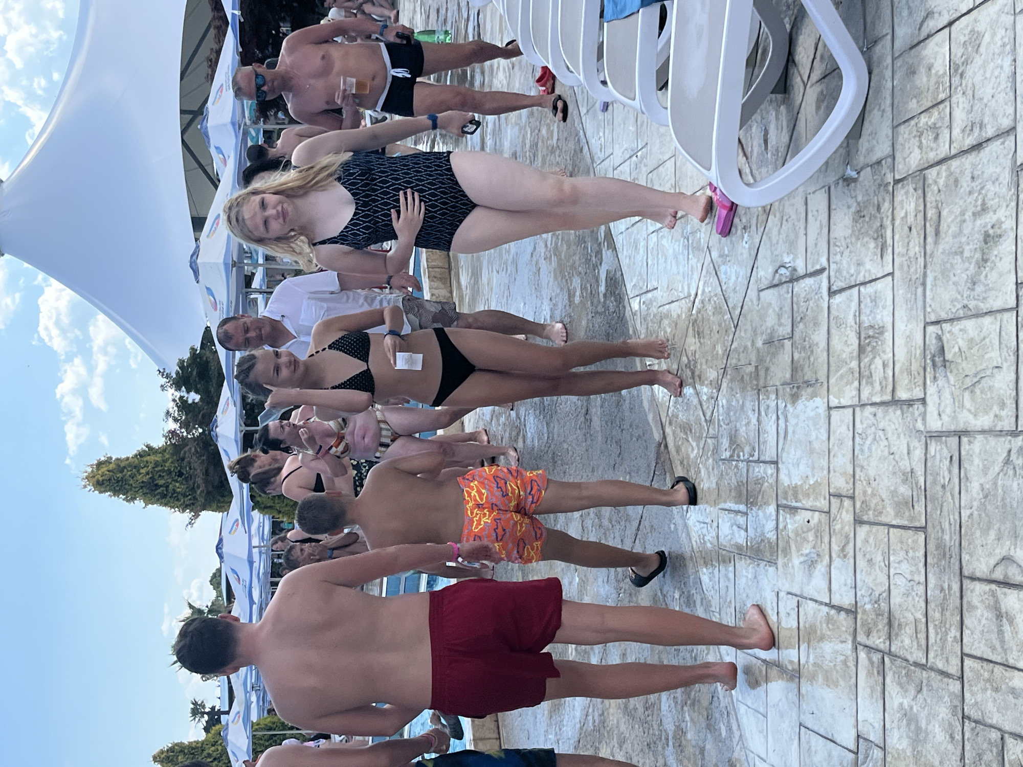 The image depicts a sunny day at a water park in Ravda, Bulgaria. A group of people, primarily dressed in swimwear, are visible walking on a wet, tiled pathway. In the foreground, a man in red swim trunks and a young boy in orange patterned swim trunks are walking away from the camera. To their right, a young woman in a black bikini and another young woman in a black patterned one-piece swimsuit are walking towards the viewer. In the background, several white umbrellas provide shade, and a large white tent structure is visible. Other individuals, including a man holding a drink, are also present. The overall atmosphere appears to be that of a leisurely summer day at a recreational facility.