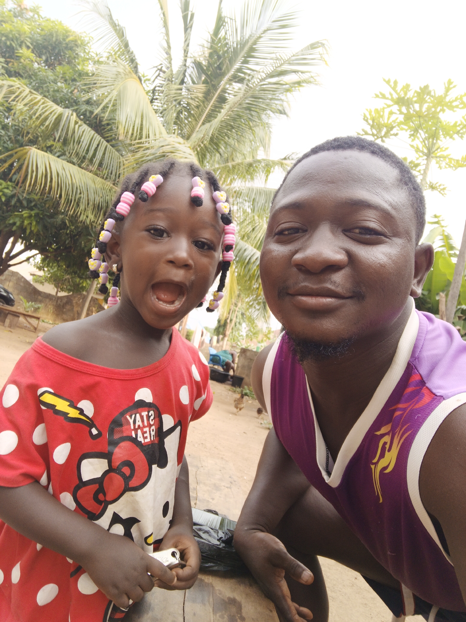 The image shows a man and a young girl in what appears to be an outdoor setting in Akwanga, Nigeria. The girl, positioned on the left, wears a red and white polka-dot shirt with a cartoon character on it and has colorful beads in her hair. Her mouth is open, suggesting she may be speaking or yelling. She also appears to be holding a metal object. The man, on the right, is wearing a purple sleeveless shirt. They are in front of trees, possibly palm trees. The location in Akwanga is suggested by the presence of tropical vegetation, and the overall atmosphere suggests a rural or semi-rural environment.