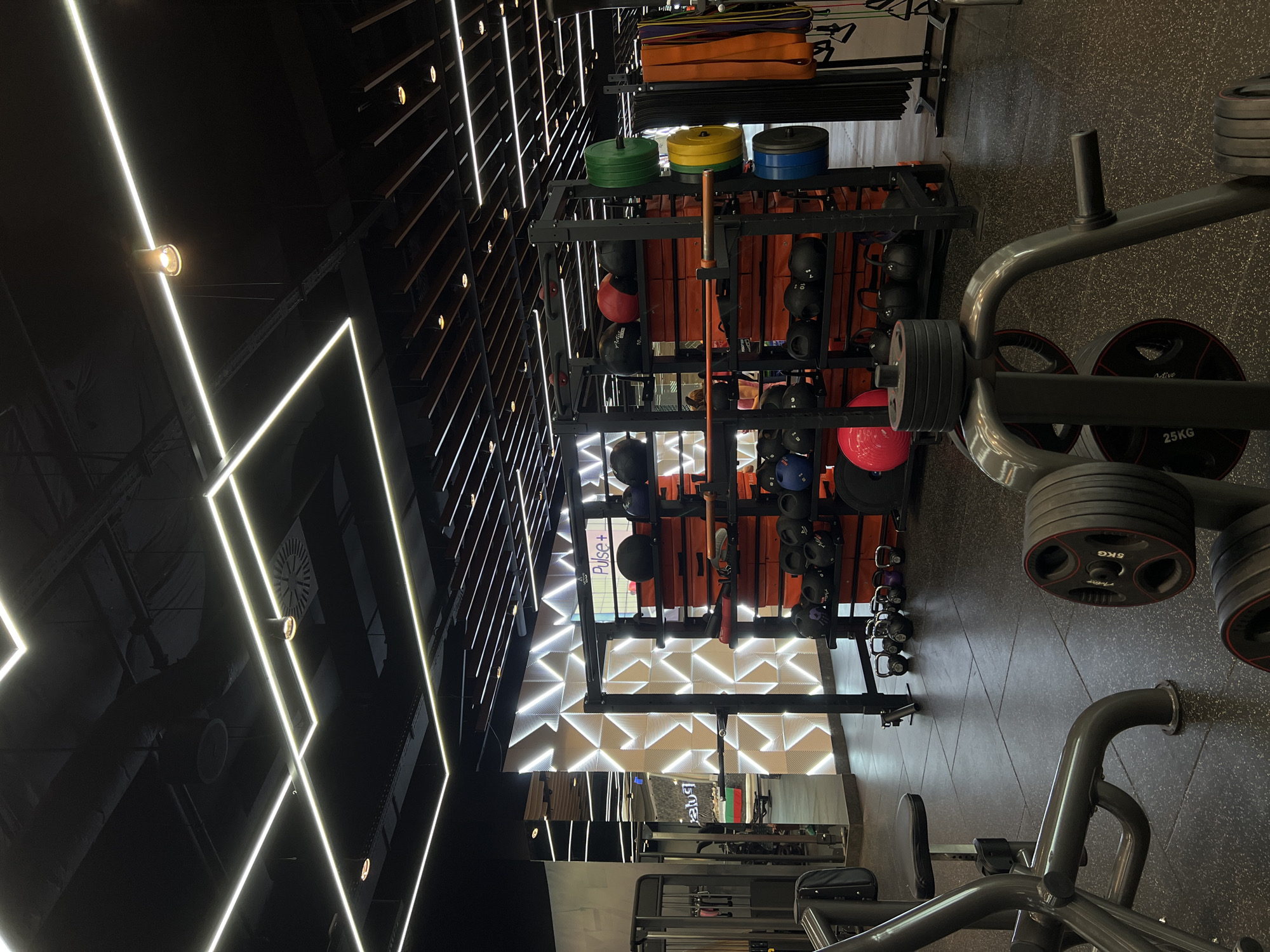 The image depicts the interior of a fitness center. Visible within the frame are weight racks stocked with dumbbells, kettlebells, medicine balls, and weight plates of varying colors and sizes. Also visible is gym equipment, including plate-loaded machines, set on a black speckled floor. The fitness center is decorated with geometric linear lighting fixtures overhead, creating a modern aesthetic. A sign with "Pulse+" is visible near the back of the center. Based on the sign and overall design elements, it is likely this fitness center is located in Burgas, Bulgaria.