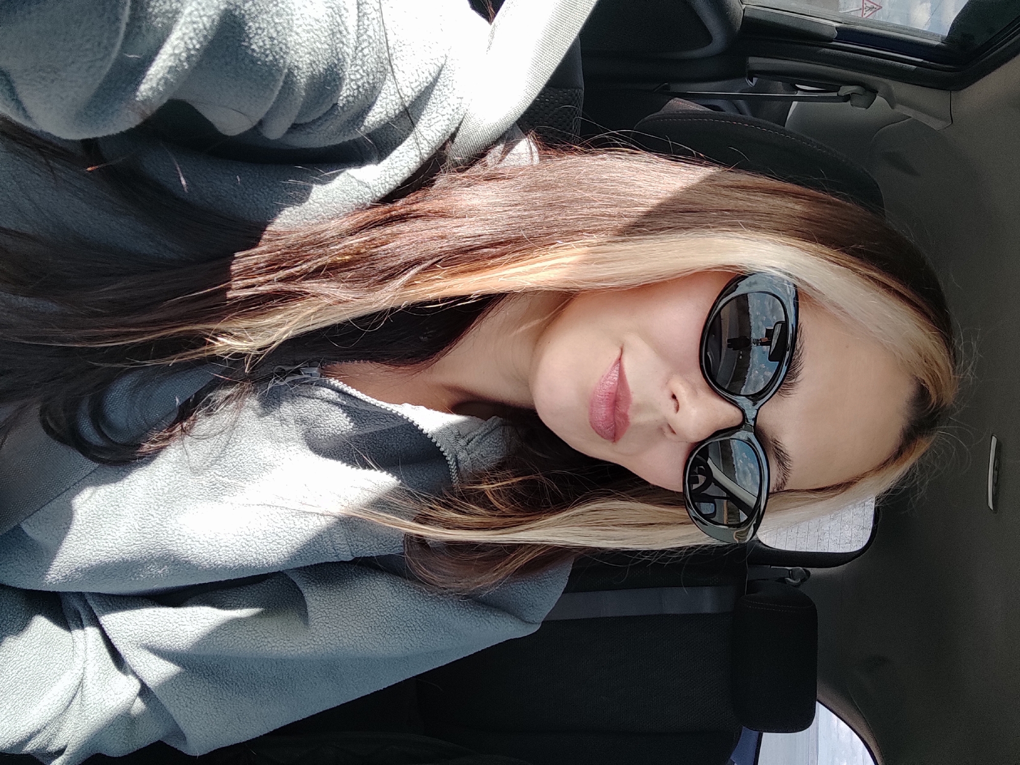 A selfie photograph shows a woman wearing sunglasses and a gray fleece jacket. She has long, brown hair with blonde streaks. The location appears to be inside a car, with a view of the car's interior, including the roof and seats. The photograph was not taken in the city of Noviy, Russia.