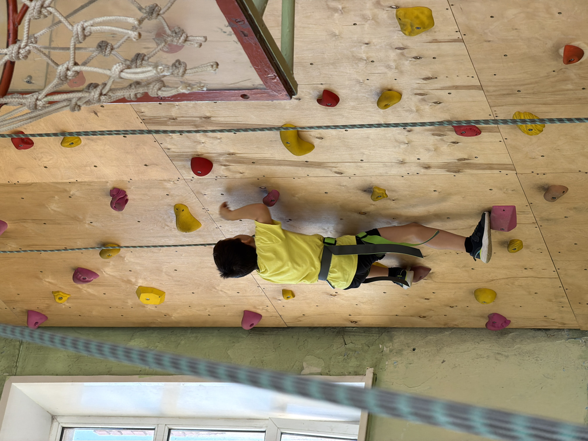 A child in a yellow shirt and climbing harness ascends a climbing wall. The wall is made of plywood and has various-colored climbing holds. A rope is visible, presumably connected to the harness for safety. A rope net hangs above. The location may be Izhevsk, Russia. A window is visible at the bottom. The scene appears to be indoors.