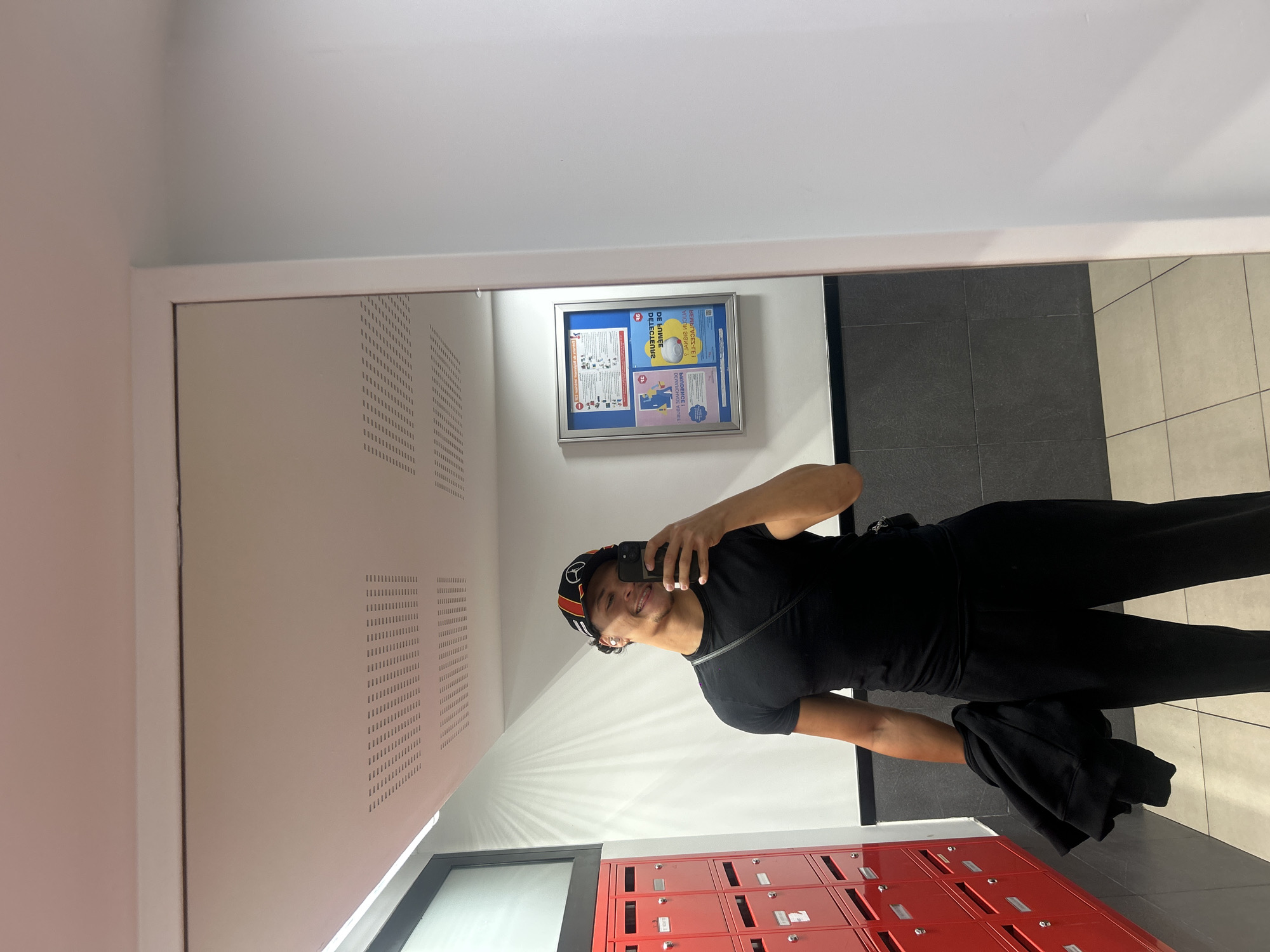 A person is captured in a mirror selfie within an indoor hallway or lobby, located in Noisy-le-Grand, France. The individual, appearing male, is seen from the waist up, holding a black smartphone to take the photo. They are smiling broadly at the camera. The person is wearing a black t-shirt, black trousers, and a black, orange, and white cap. A black jacket or sweatshirt is casually held in their left hand.
The setting is a well-lit space with white walls and ceiling. To the left of the person (in the reflection), there are perforated panels or ventilation grates on the wall. Directly below the person, a bank of red mailboxes or lockers with silver locks is visible. In the background, behind the person's reflection, there is a framed blue bulletin board displaying various notices and diagrams, including text that appears reversed due to the mirror, with fragments like "DE L'ANNEE" and "JANDI KUDUA" partially legible. The floor is tiled, featuring dark grey tiles on one side and lighter, possibly beige, tiles on the other. The overall impression is a modern, communal area within a building.