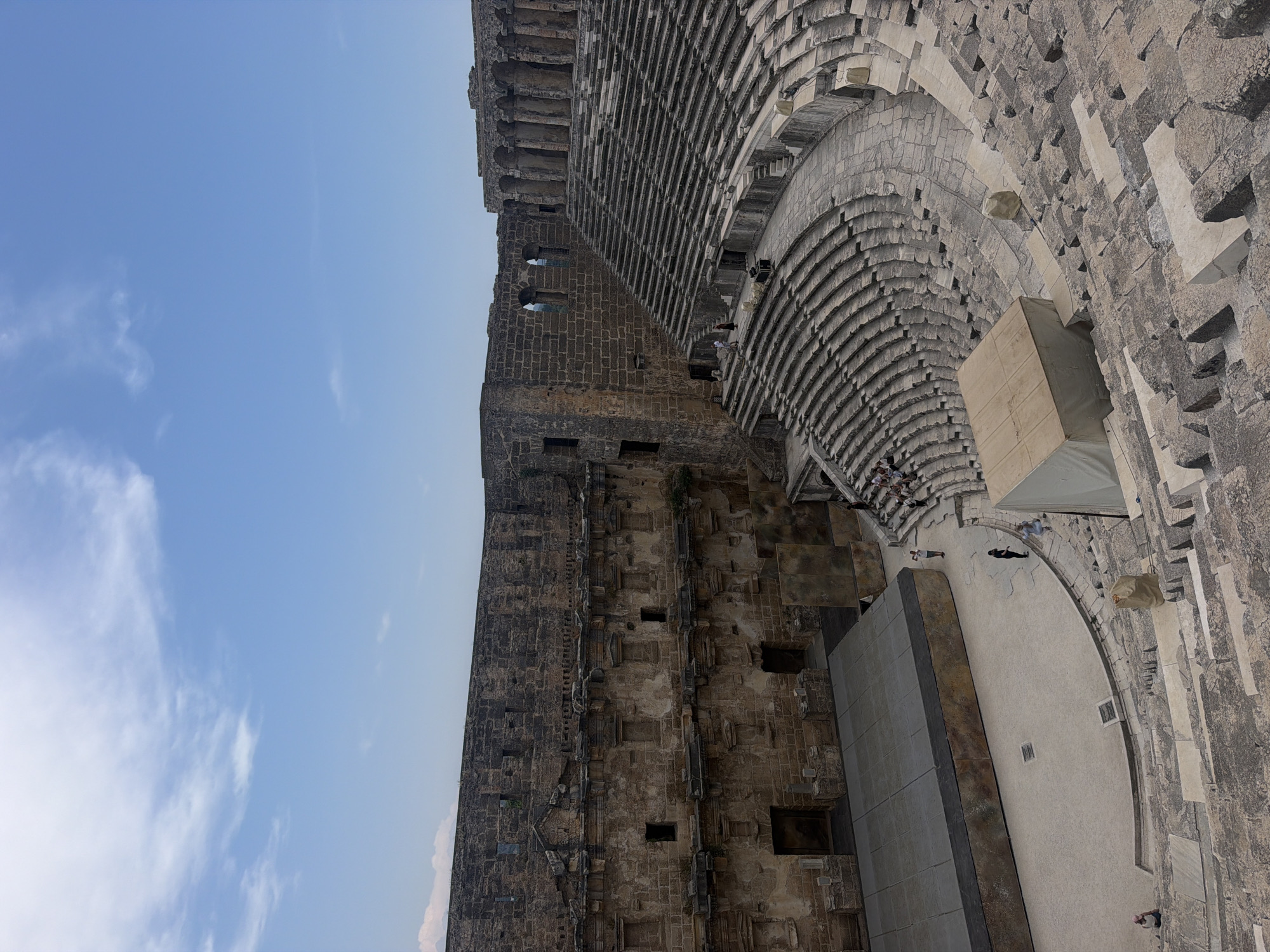 The image shows the Aspendos Theater, a well-preserved Roman theater located in Serik, Turkey. The theater is made of stone and has a semicircular seating area that curves around a stage. There are several people visible in the image, most of whom are seated in the theater or walking on the stage. The sky is blue with some clouds. The Aspendos Theater is a popular tourist attraction and is known for its excellent acoustics.