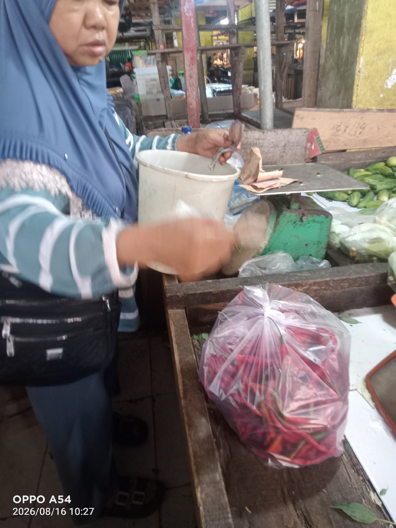 The image depicts a woman in a blue hijab and a striped shirt at a market stall, possibly in Jambi, Indonesia. She is using a scoop to transfer red chilies from a plastic bag into a white bucket. In the background, a green scale is visible, along with other vegetables and market stalls. The overall scene suggests a transaction or preparation for sale of produce at a local market. The time and date stamp on the image indicate it was taken on August 16, 2026, at 10:27.