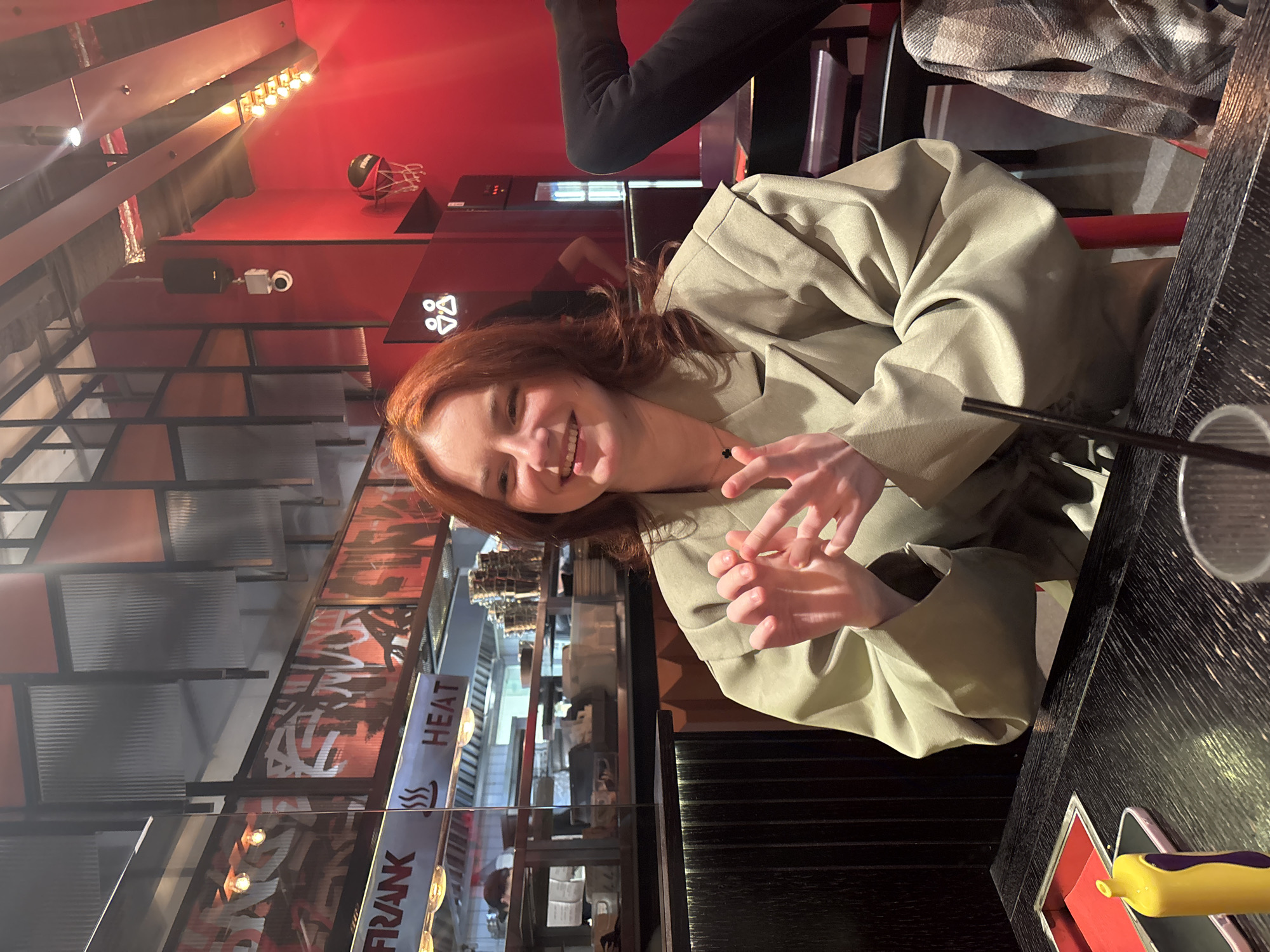 A woman with red hair is sitting at a table in a restaurant in the city of Unknown, Russia. She is wearing a light green jacket. There is a glass and a yellow object on the table. The restaurant has red walls and metal structures with signs that read "FRANK" and "HEAT". There is a basketball hoop on the wall. Another person is partially visible in the background.