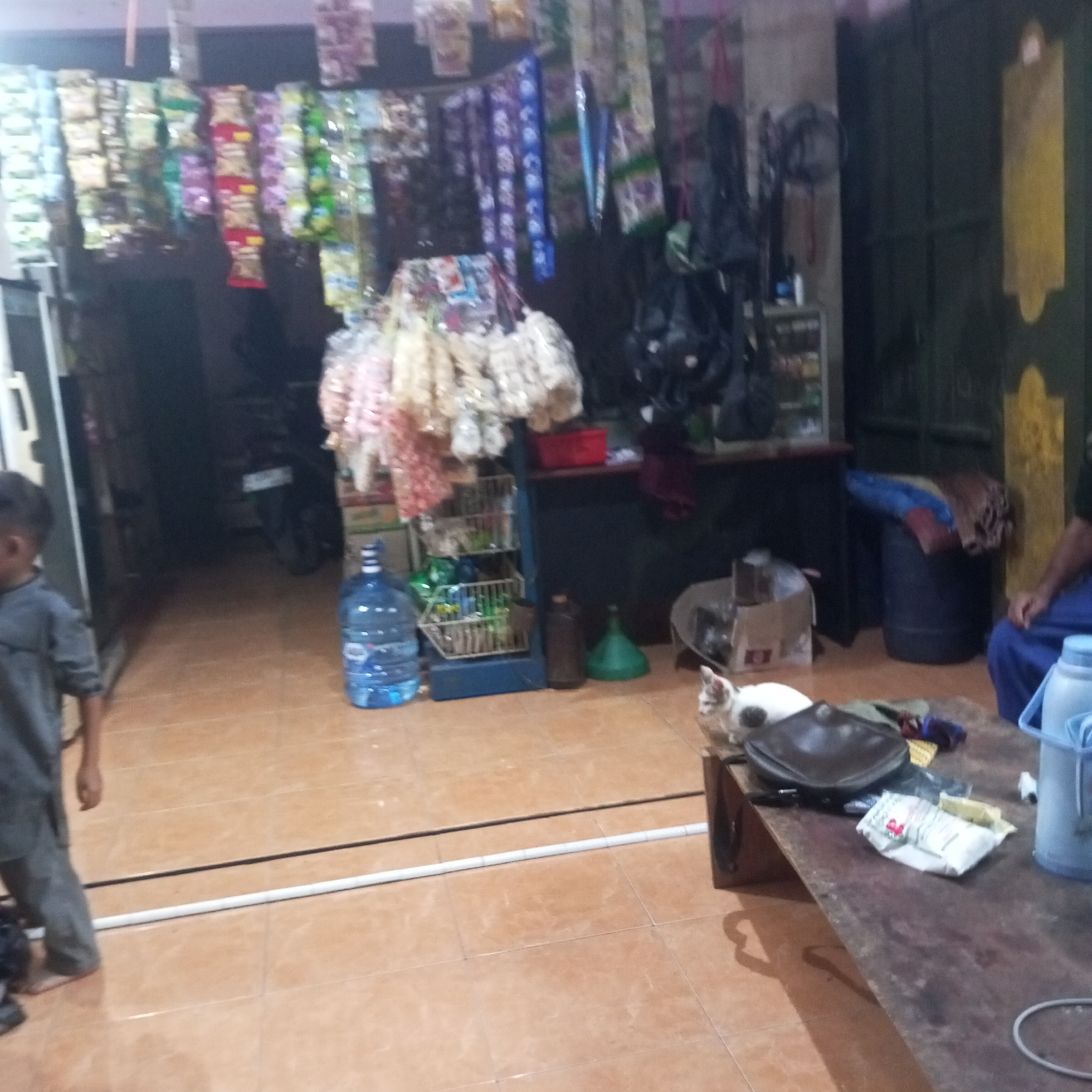 The image depicts the interior of a small shop, possibly a convenience store or warung, located in Cibatuireng, Indonesia. The shop is filled with various goods displayed on shelves and hanging from the ceiling, including packaged snacks and other merchandise. A young boy in a gray outfit stands on the left side of the image, facing towards the right. In the background, a motorcycle is parked inside the shop. On the right side, a wooden table is present, with a cat resting on it. A person wearing blue pants is partially visible sitting to the right of the table. The floor is tiled in a reddish-brown color. The overall impression is that of a functional, modestly stocked shop.