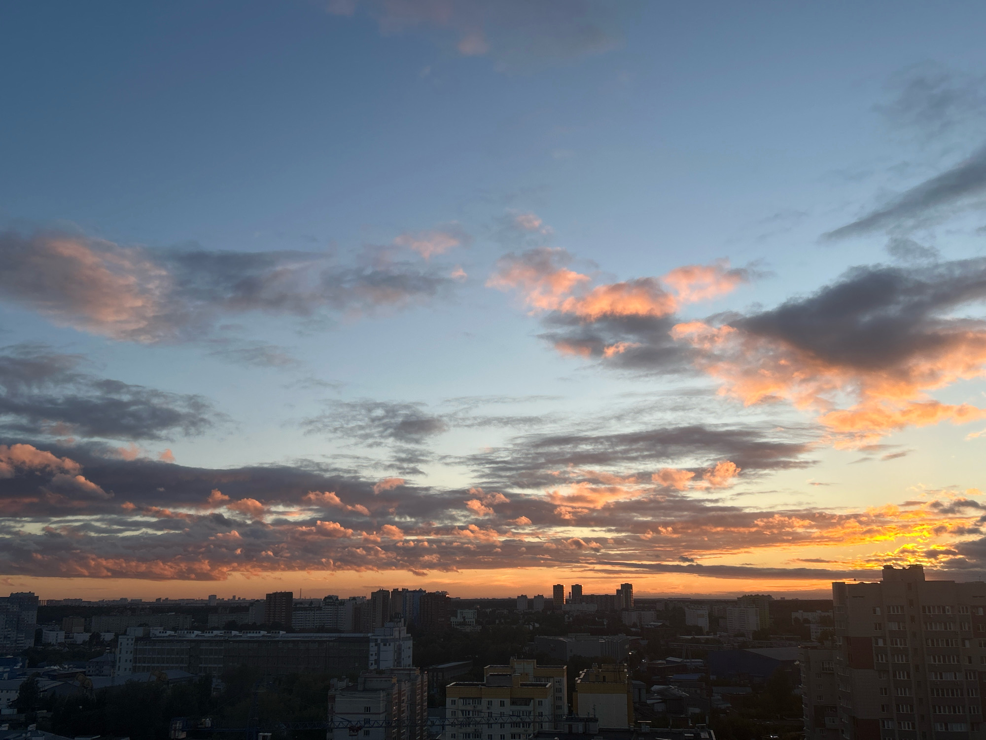 The image depicts a panoramic view of the city of Ivanteyevka, Russia, during what appears to be a sunset. The sky is a gradient of blues and oranges, with clouds illuminated by the setting sun, casting a warm glow. Silhouetted against the vibrant sky are numerous apartment buildings and other structures characteristic of a city skyline. The architecture suggests a dense urban environment with multi-story buildings. The overall scene is one of a developing twilight over an inhabited area. There are no visible people or specific actions taking place, with the focus being on the cityscape and the atmospheric conditions of the sky.