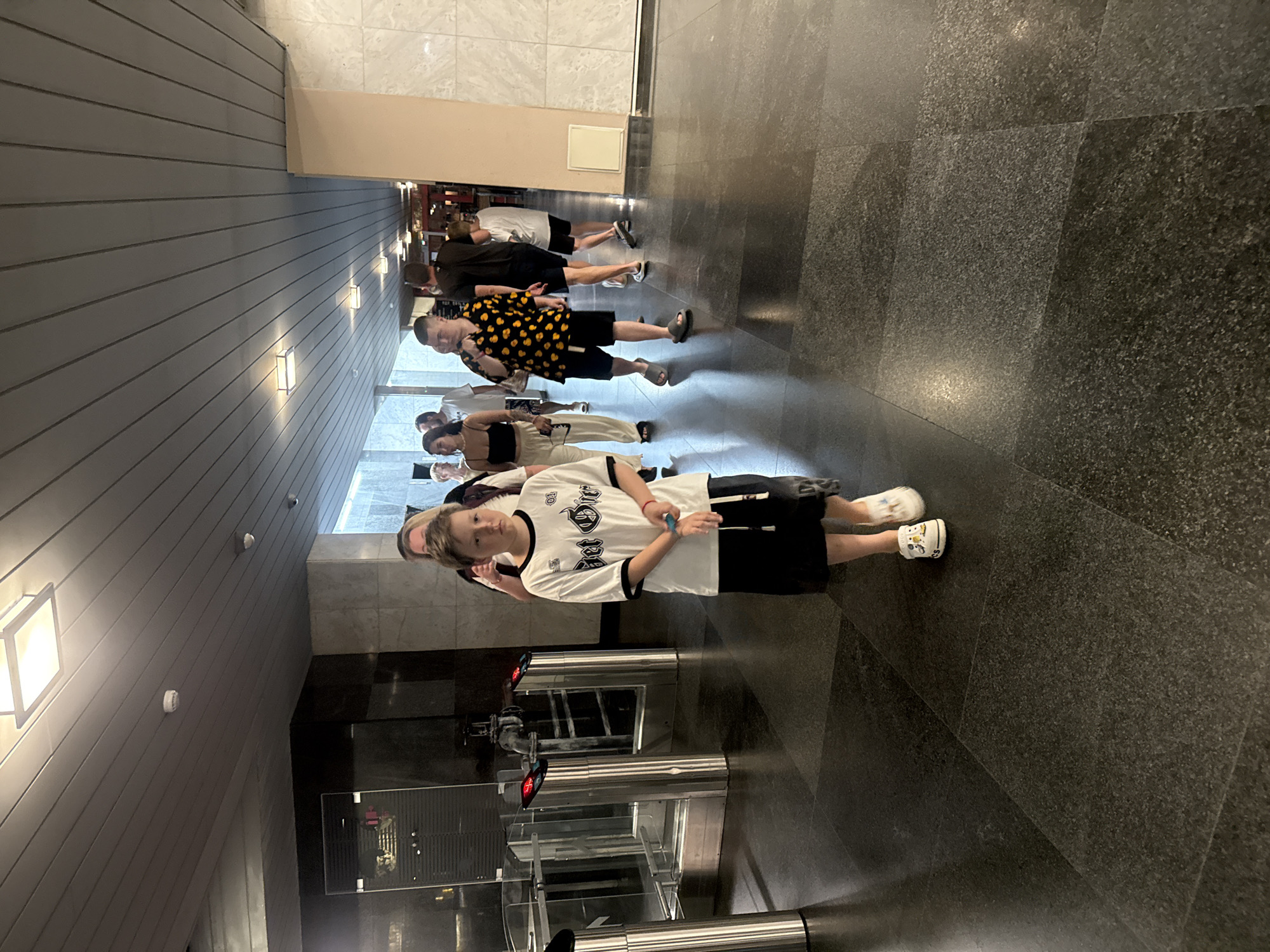 A boy in a white t-shirt and black shorts stands in the foreground of a brightly lit indoor hallway. He is looking directly at the camera with a neutral expression. Behind him, a group of people, including men, women, and children, are walking in the same direction. They appear to be in a public space, possibly a museum or entertainment venue, indicated by the turnstiles on the left side of the image. The hallway is modern, with marble walls and a tiled floor. The lighting is from overhead fluorescent fixtures. The scene appears to be taking place in Yalta, Ukraine.