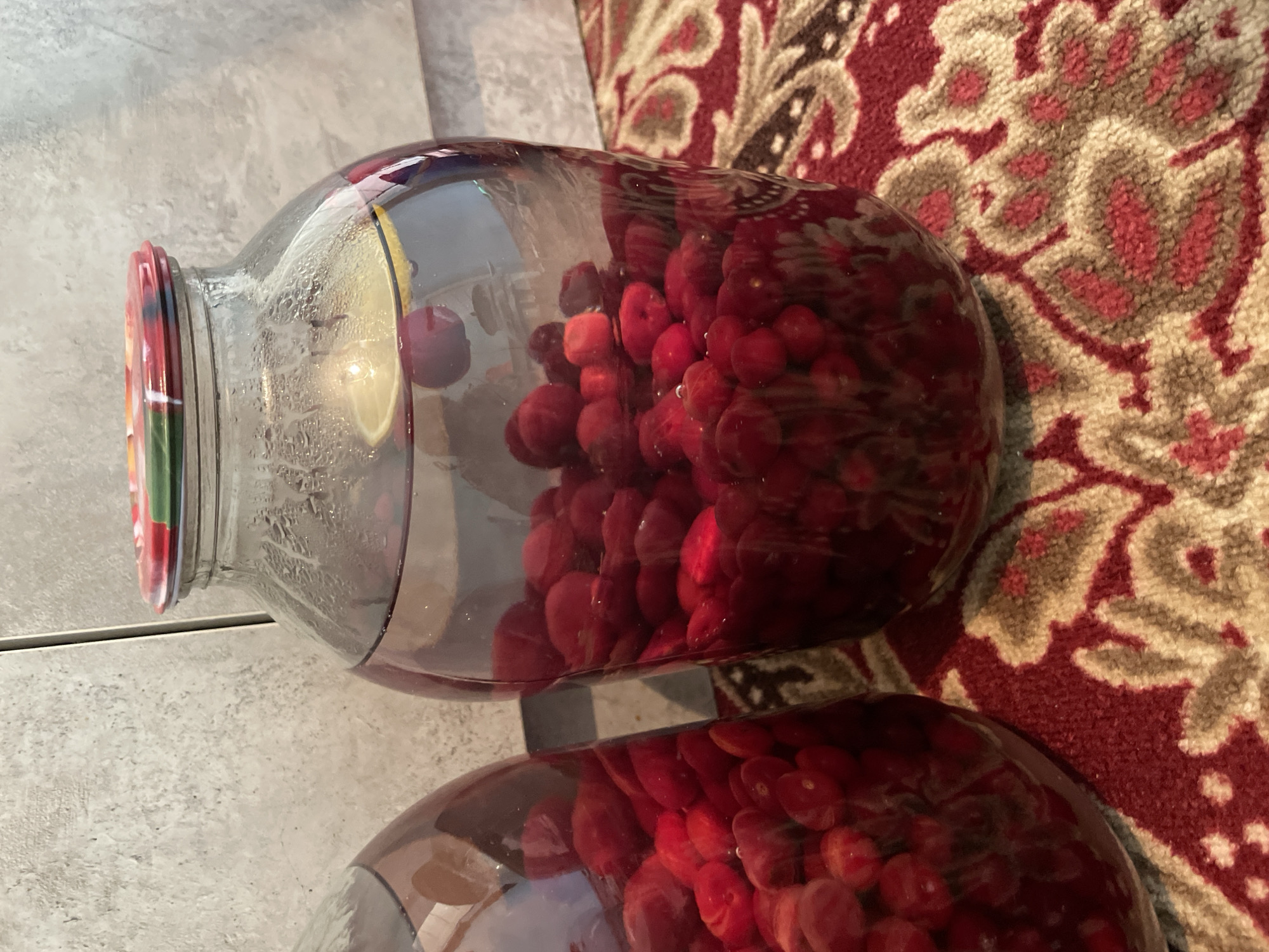 In an indoor setting, two large glass jars filled with preserved red berries, possibly cherries or cranberries, are positioned on a red and beige patterned rug. The jar in the foreground is partially filled with berries and clear liquid, with a slice of lemon floating near the top. The lid of this jar is colorful, with a pattern of red, green, and yellow. The second jar, visible on the left, is also filled with berries and liquid. The background features a textured gray wall, possibly tile or concrete. There are no people visible in the image. The scene is not specific to the city of Bujum, Kyrgyzstan, as no identifying landmarks or contextual clues are present.