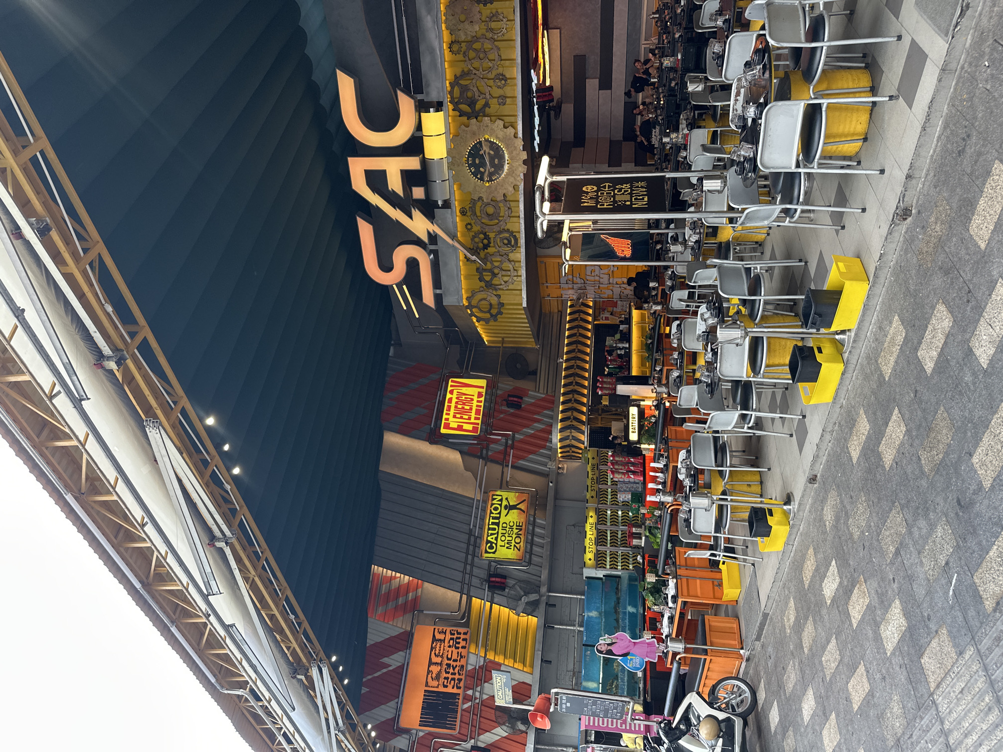 This media file depicts an outdoor seating area of a restaurant or bar with a distinct industrial-themed decor. The scene is viewed from a low angle, looking upwards and across the tables and chairs arranged along a sidewalk. The restaurant facade is decorated with large metallic gears and exposed metal elements, contributing to its industrial aesthetic. Prominently displayed are several signs, including "SAC," "CAUTION LOUD MUSIC ZONE," and "ENERGY," suggesting the establishment's ambiance and offerings.
The seating area consists of numerous tables with chairs, many of which have yellow bases or accents. Some chairs are occupied by patrons, though they are not the primary focus. In the foreground, there are a few motorcycles parked, one with a helmet on the seat. The ground is paved with a checkered pattern of gray and beige tiles. The time of day appears to be daytime, with bright lighting, possibly mid-afternoon, given the shadows. The setting seems to be a commercial street in Thủ Đức, Vietnam, characterized by the architecture and the general urban environment. The overall mood is lively and casual, indicating a popular spot for dining or socializing.
