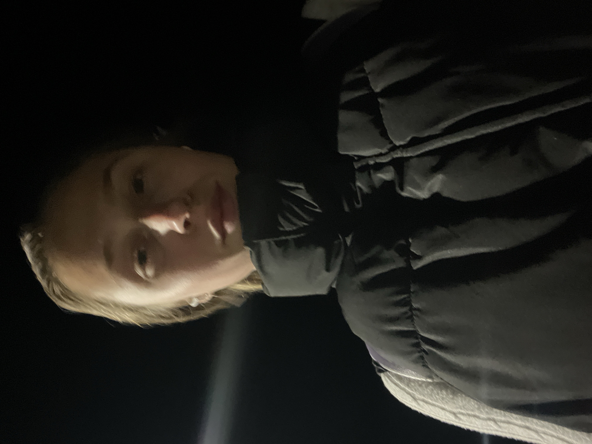 A dimly lit, angled selfie of a blonde woman wearing a black puffy jacket with a high collar. The background is primarily black, suggesting nighttime. The woman's face is illuminated by a light source, likely from below, accentuating her features. The woman's left eye is visible as well as her nose and mouth. Her short blonde hair is visible on the left side of the frame. The environment is indistinct, but the darkness and absence of clear landmarks suggest an outdoor location. The location, Budva, Montenegro, cannot be verified due to the absence of recognizable landmarks in the image.