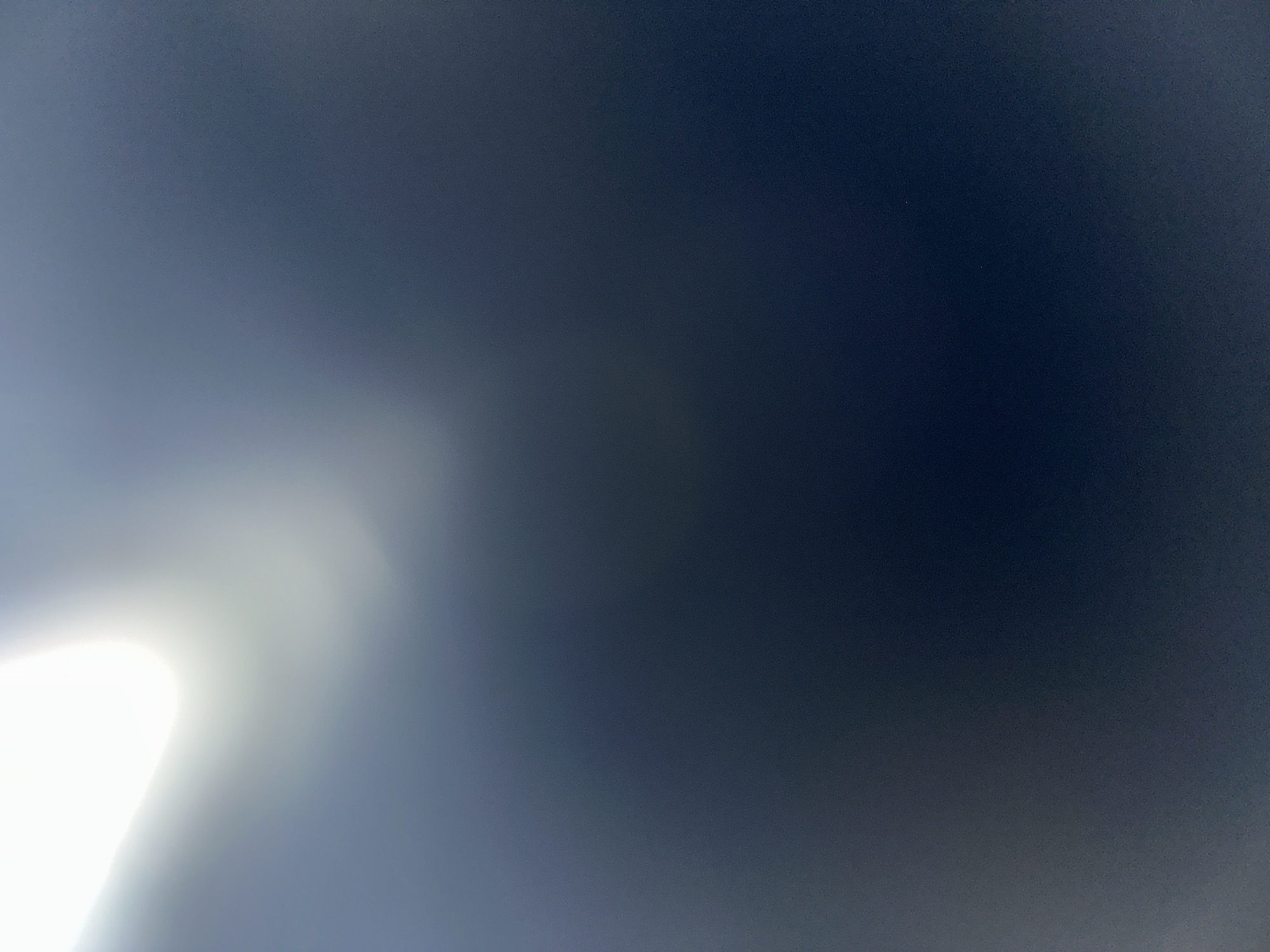 This is a photograph that appears to be taken from a low angle, looking upwards towards a sky that transitions from a bright, luminous white on the left side of the frame to a deep, dark blue or black on the right. The bright area suggests a strong light source, possibly the sun, which is out of frame or partially obscured. There are no discernible people, objects, or specific scene elements. The image is abstract and lacks clear subject matter. The lighting creates a gradient effect, with the brightest part likely representing a direct light source and the darker portion representing the sky further away or in shadow. There is no visible text. Given the context of Belgrade, Serbia, and the nature of the lighting, it's possible this photo was taken outdoors on a clear day, perhaps with the camera pointed towards the sun or a very bright artificial light source. The time of day is difficult to ascertain precisely due to the abstract nature of the image, but the bright white suggests a time when the sun is high or intensely bright. There are no indications of weather conditions beyond the presence of light.