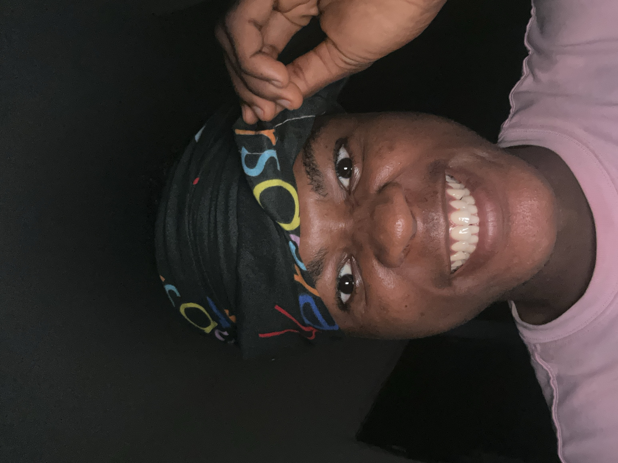 A close-up, low-angle shot captures a Black individual from the chest up, smiling broadly. They are wearing a pink t-shirt and a black bandana adorned with colorful letters. The individual's right hand is raised, holding the edge of the bandana near their temple. The lighting is dim, casting a shadow behind them, suggesting an indoor setting at night. The background is dark and indistinct. This image was taken in Ihiagwa, Nigeria.