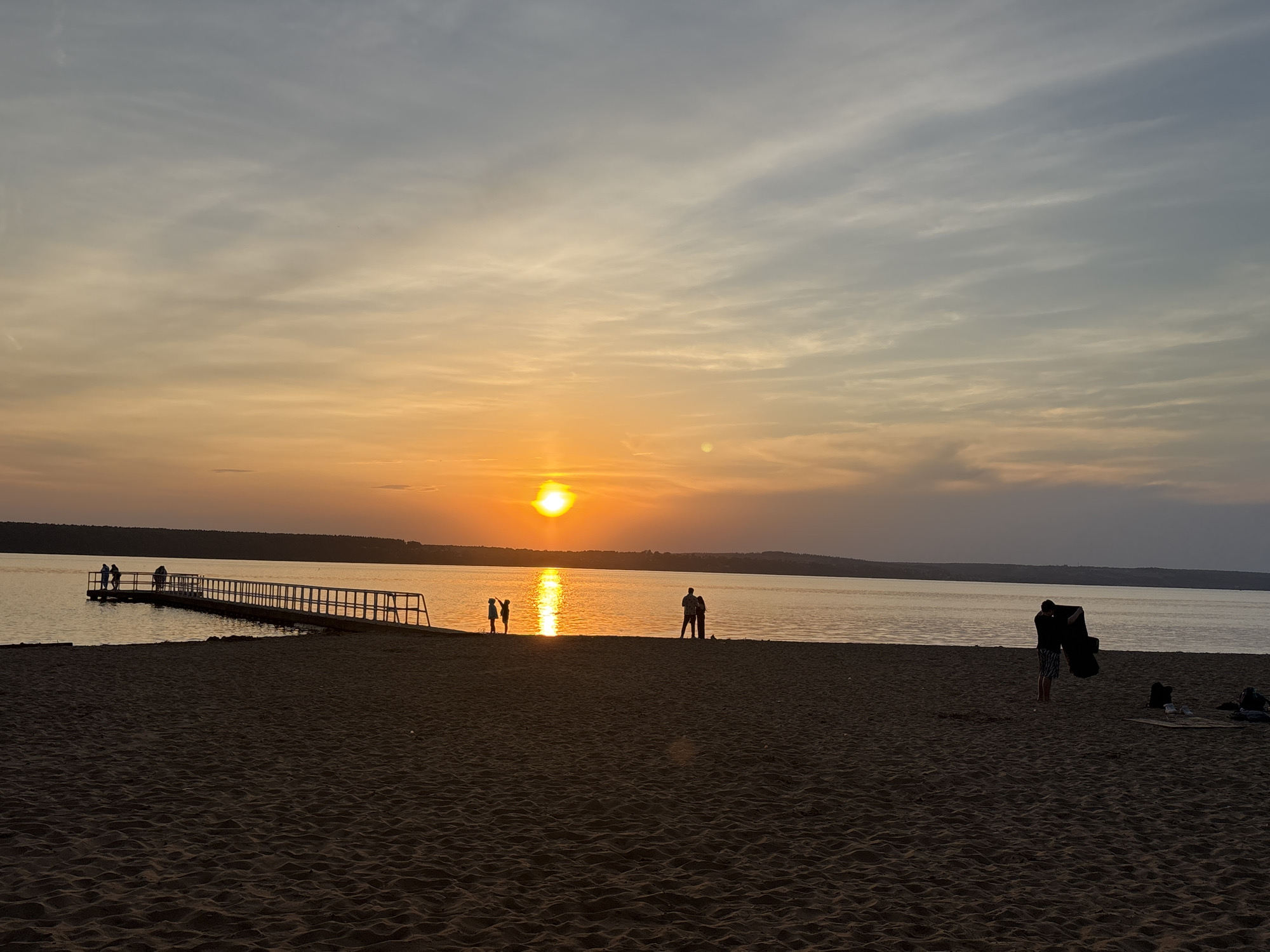The image depicts a sunset scene at a beach in Perm, Russia. The sun is setting over a body of water, with its reflection visible on the surface. A long pier extends into the water on the left side of the frame, with several people standing on it. In the water, a few figures are silhouetted against the setting sun. On the beach in the foreground, individuals are seen standing, sitting, or interacting. One person stands with a dark object in their hands. The beach consists of sand, and the sky transitions from orange near the horizon to a lighter, cloudier blue above. The treeline is visible in the background across the water.