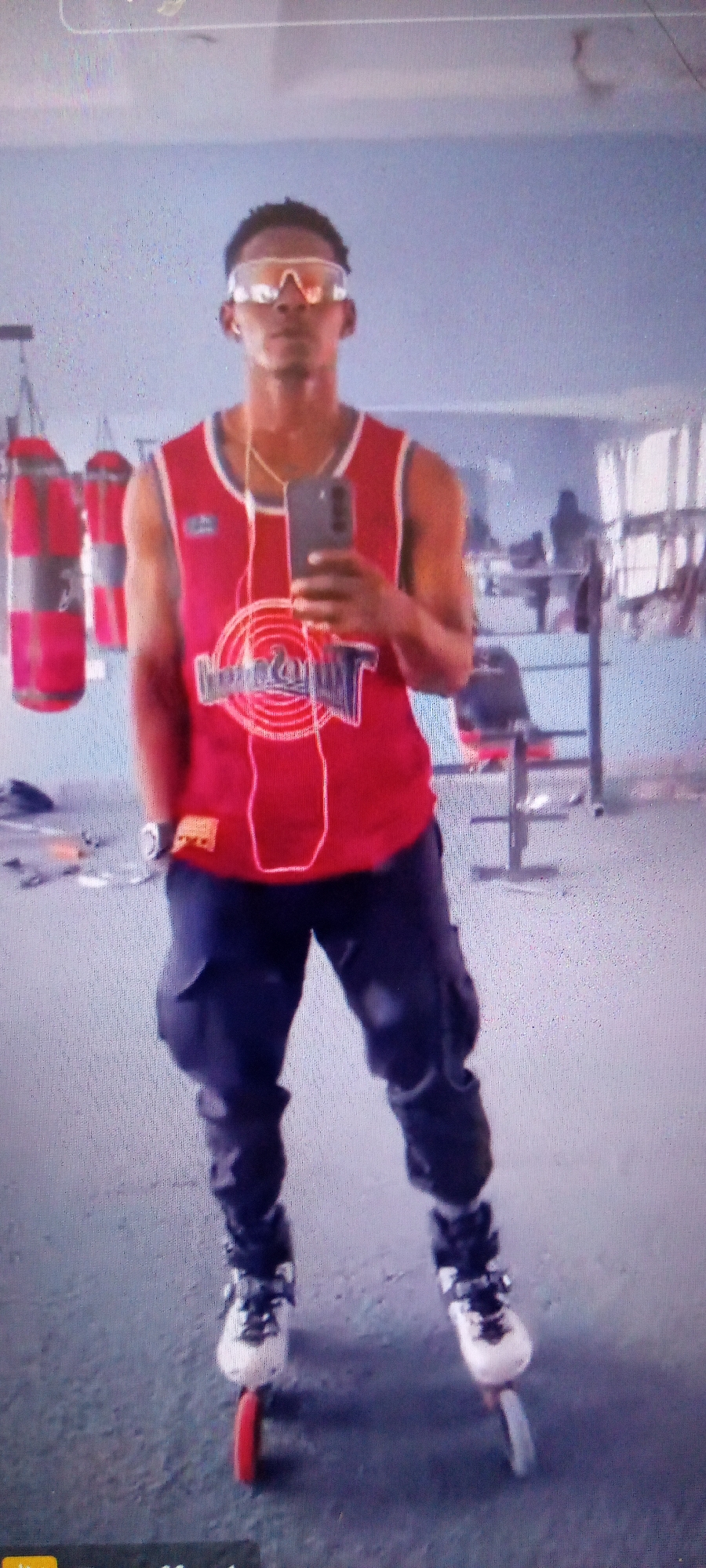 A young man stands centered in an indoor gymnasium, appearing to take a selfie. He wears clear wraparound sunglasses, small earbuds, and a gold-colored chain around his neck. His dark hair is short and styled. He is dressed in a red sleeveless jersey, featuring a prominent circular "Space Jam" logo with Bugs Bunny on the front, and dark cargo pants. On his feet are white and black inline skates, with a red wheel clearly visible on the left skate.
He holds a smartphone in his right hand, pointed towards himself, suggesting he is capturing a self-portrait, with his reflection visible on the phone's screen. The background of the image displays typical gym equipment, including several red punching bags hanging on the left side, and various weightlifting benches and racks scattered across the room. The floor is a dark gray, likely rubber, and the walls are a light, muted blue or gray. The lighting appears artificial, characteristic of an indoor sports facility. The scene is set in Akwanga, Nigeria.
