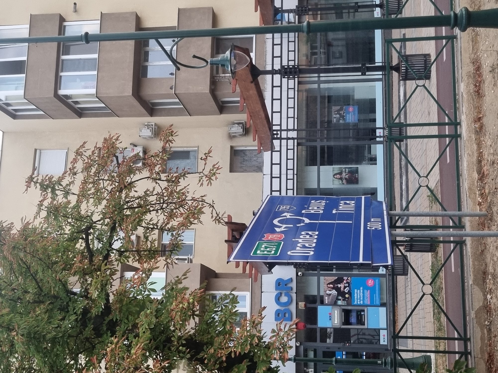 The image shows a street scene in Salonta, Romania, with a focus on a blue road sign with white text. The sign indicates directions to "Oradea" and "Beius," with "Tinca" listed below. European route E671 and Romanian national road 79 are also marked on the sign. The sign indicates that Oradea is straight ahead, Beius is to the right, and Tinca is also to the right. A distance marker of "500 m" is present below the directional information. In the background, a modern building with balconies and windows is visible. Part of a BCR (Romanian Commercial Bank) branch with an ATM and posters is also visible to the left of the sign. A tree with green and brown leaves partially obscures the upper portion of the image. A green lamppost stands to the right of the sign. A decorative metal fence runs along the sidewalk in the foreground. There are no people clearly visible in the image.