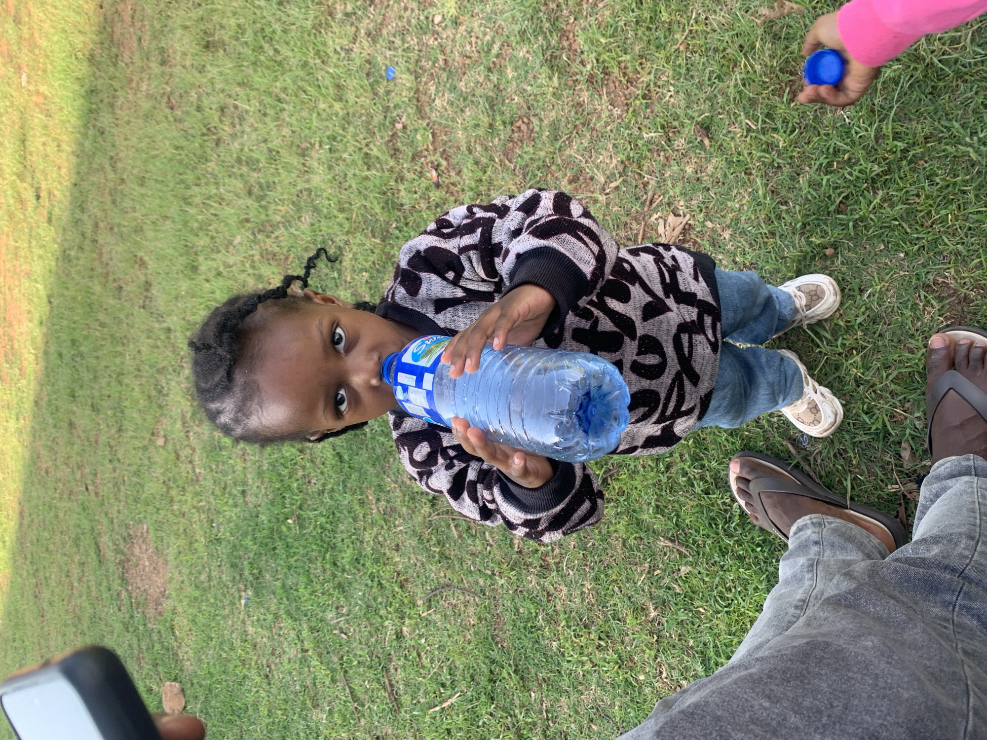 A young Black child with braided hair stands on a grassy area, holding a partially full bottle of water to their mouth and drinking. The child is wearing a patterned sweater and blue jeans. Their feet are clad in white sneakers with a beige logo pattern. To the left of the child, a portion of an adult's legs and feet are visible, wearing denim jeans and brown flip-flops. Another adult's arm and hand, wearing a pink long-sleeved garment, reaches into the frame from the right, holding a blue bottle cap. The background consists of green grass with some dry leaves and twigs scattered on it. The scene appears to be an outdoor public space in Jos, Nigeria, possibly a park or a recreational area. The lighting suggests it is daytime.