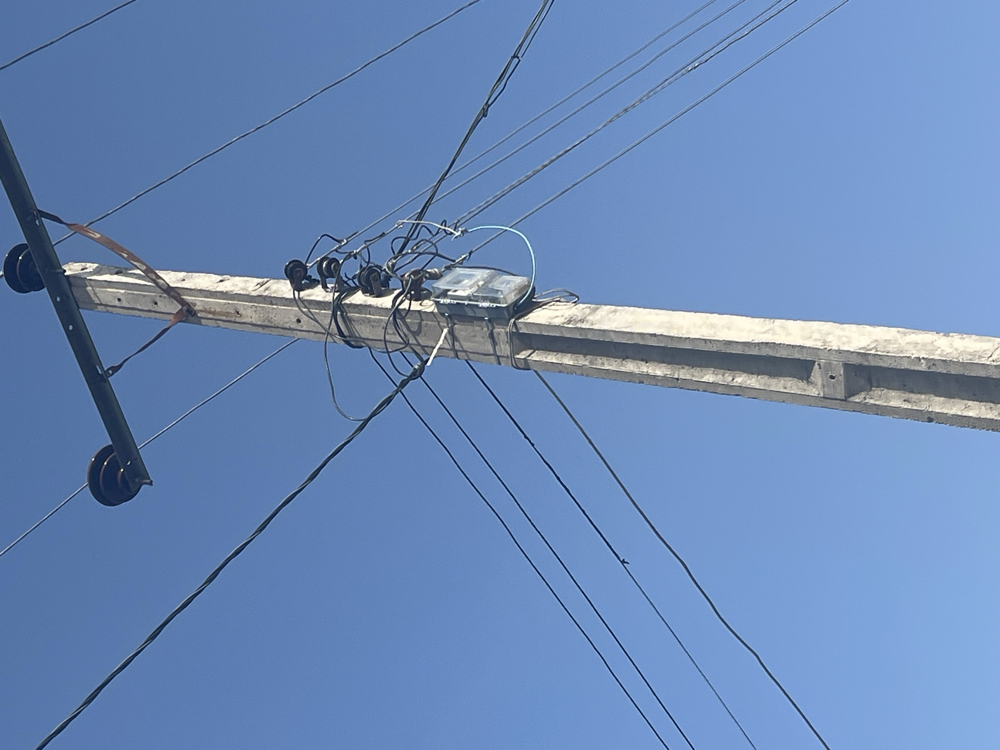 The image depicts a concrete utility pole against a clear blue sky. The pole is equipped with a horizontal crossbar supporting dark, circular insulators. Several wires are strung from these insulators, extending across the frame. Attached to the pole, midway up, is a gray electrical meter housed in a clear plastic casing. Numerous wires converge and connect to this meter, with one prominent light blue wire looping from the meter. The scene is characteristic of urban infrastructure in Jalingo, Nigeria, where such poles and meters are common for power distribution.