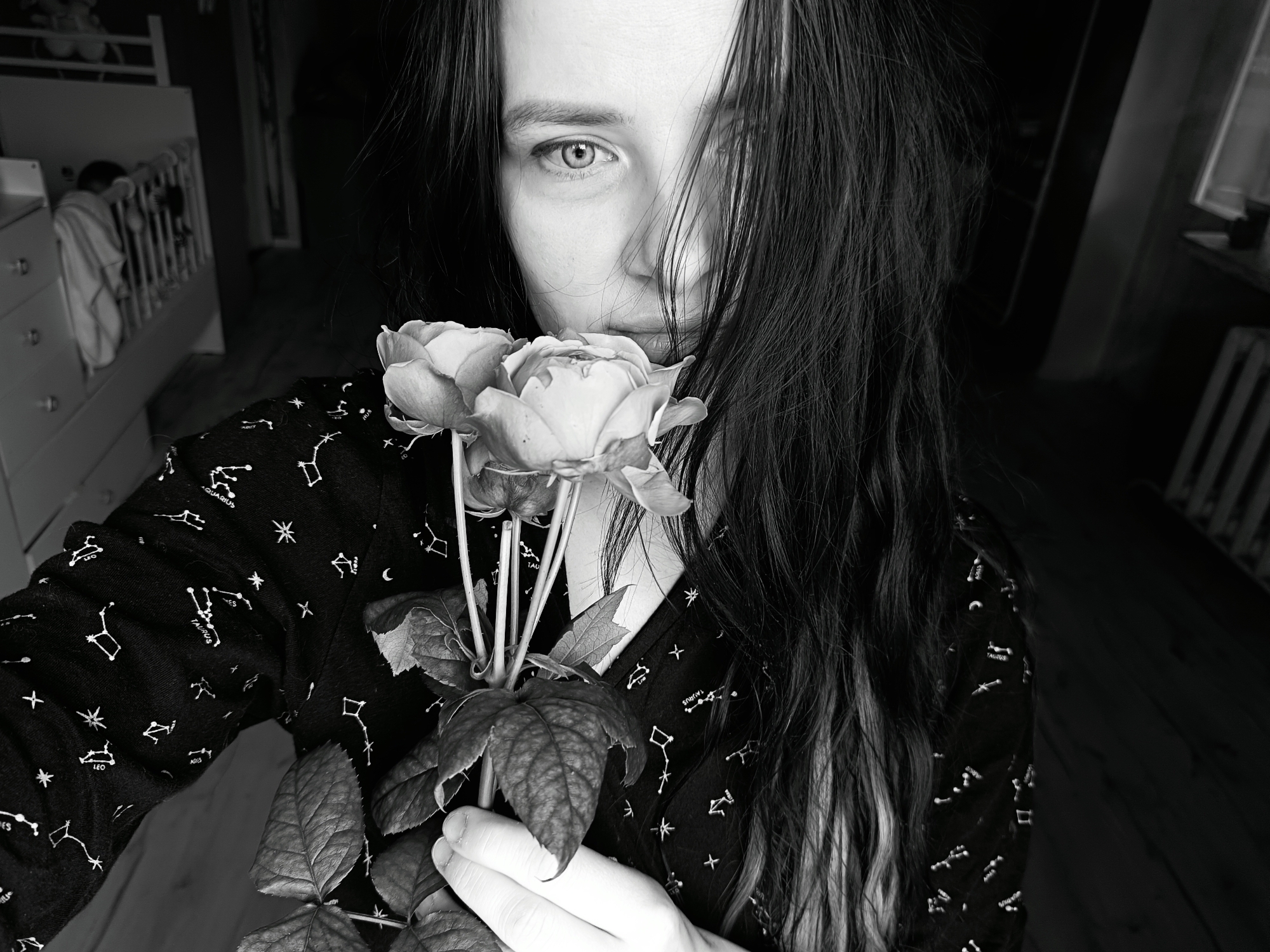 The black and white photograph captures a close-up, intimate portrait of a person holding a bunch of roses. The subject, likely female, has long, dark, slightly wavy hair that partially obscures her face, with one light-colored eye gazing directly at the viewer. Her nose and mouth are subtly visible behind the flowers. She wears a dark, long-sleeved shirt adorned with white patterns of constellations, including identifiable text such as "AQUARIUS," "LEO," and "TAURUS."
The person holds two prominent rose blooms, along with several leaves, close to her face. Her hand, with visible knuckles and fingers, supports the stems.
In the blurred background to the left, a white wooden crib or cot is visible, with a figure (possibly a baby or child) partially discernible inside, draped in light fabric. Next to the crib is a white dresser or cabinet. The right side of the background is darker, showing hints of an interior space. The setting appears to be indoors, likely a home, possibly a bedroom or nursery in Rusalevka, Russia.
The lighting is soft and somewhat dim, contributing to the photograph's introspective and artistic mood. The black and white conversion emphasizes textures, contrasts, and the subject's direct gaze. No specific time of day or weather can be definitively determined from the image, but the indoor setting and lighting suggest a calm, perhaps quiet, moment.