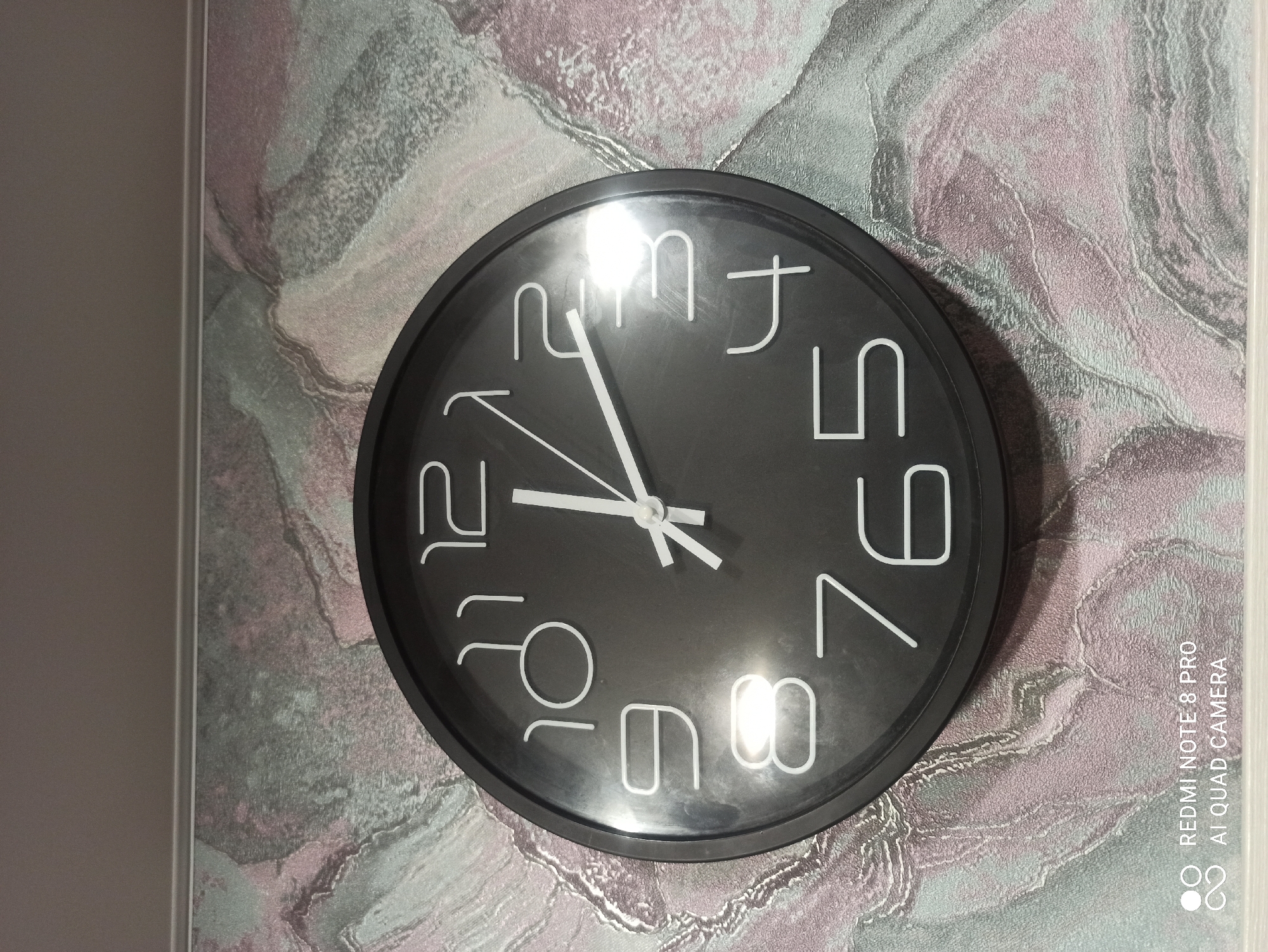 A black analog clock is mounted on a textured wallpaper with pink and grey marbled patterns. The clock face is black with white numbers and white hands. The hour hand is pointing at the 12, and the minute hand is pointing just past the 12, indicating the time is approximately 12:02. The image is taken from a slightly elevated angle, looking down at the clock. The bottom left corner of the image contains a watermark that reads "REDMI NOTE 8 PRO AI QUAD CAMERA". There are no people or any other objects visible in the frame. The location of the image is Kugesi, Russia.