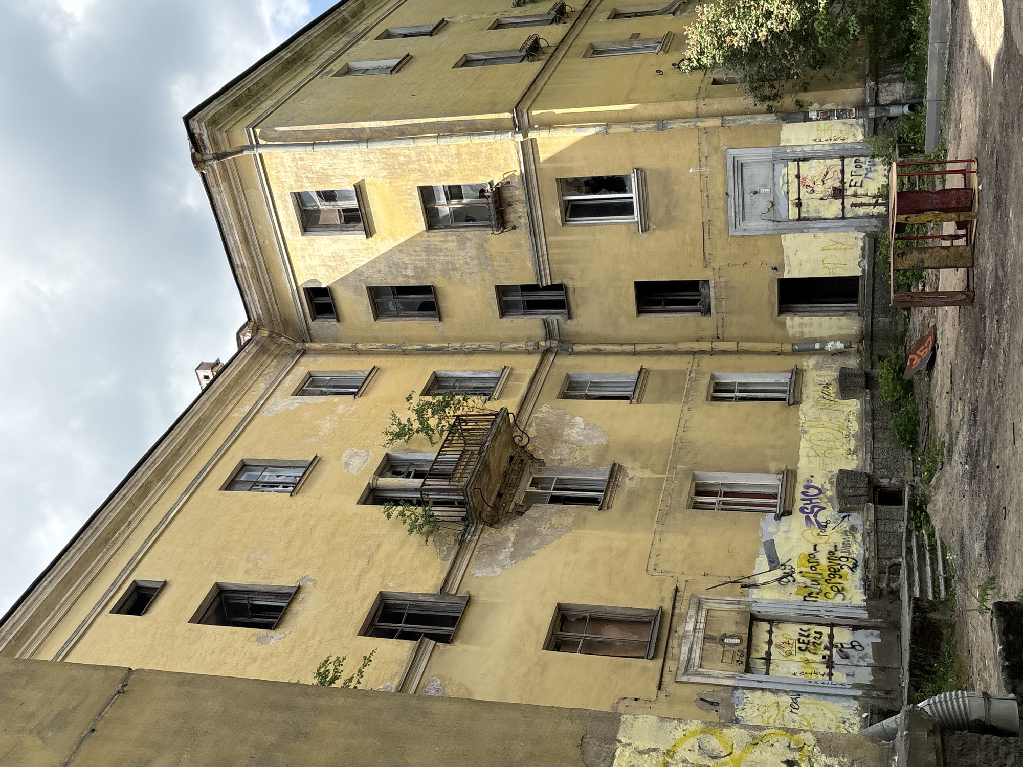 The image depicts a weathered, yellow apartment building in Saint Petersburg, Russia, likely from the Soviet era, given its architectural style and condition. The building shows signs of neglect and decay, with peeling paint, broken windows, and graffiti adorning its facade. A small, rusted balcony with overgrown plants juts out from the second floor. In the foreground, a dilapidated playground structure, consisting of metal posts and wooden planks, sits on a patch of uneven dirt. The overall scene conveys a sense of abandonment and urban decay.