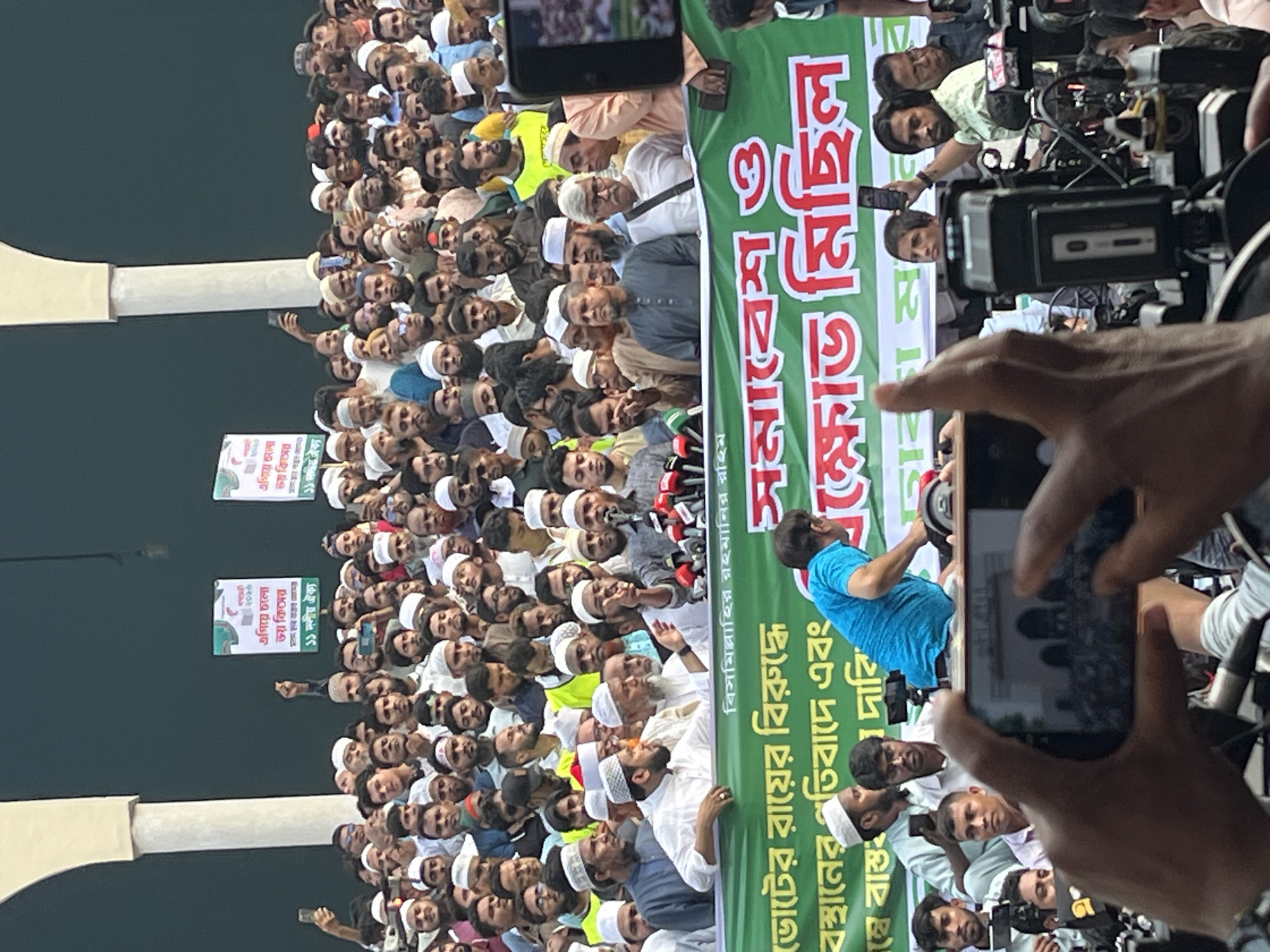 A large crowd of people, predominantly men, gathers in what appears to be a public space in Dhaka, Bangladesh. Many individuals wear traditional Muslim attire, including skullcaps. They are positioned near a green banner with Bengali script and Arabic phrase (Bismillah ir-Rahman ir-Rahim, "In the name of God, the Most Gracious, the Most Merciful"). A person in a blue shirt is partially visible, seemingly addressing the crowd from behind the banner. The crowd is being recorded by multiple cameras and smartphones. Two vertically oriented paper signs are partially visible. The architectural element in the background suggests the presence of a significant structure, potentially a mosque or gate.