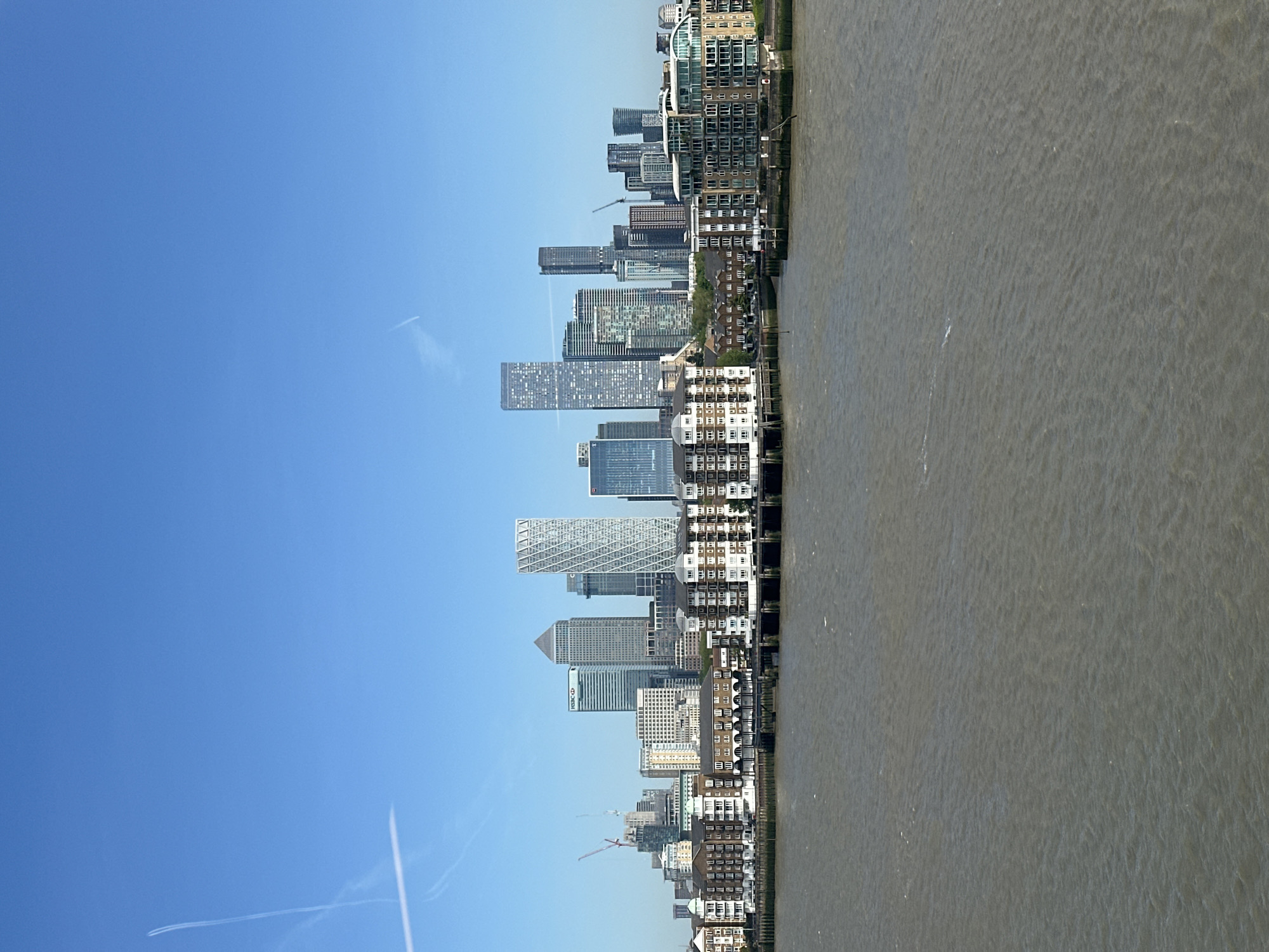 This image captures a panoramic view of a modern urban skyline adjacent to a wide river under a clear blue sky. The photograph is taken from an elevated perspective, looking across the water towards the city.
The upper portion of the frame is dominated by a bright, clear blue sky, featuring several faint white contrails from airplanes, suggesting a busy air traffic corridor. No clouds are visible.
The central part of the image showcases a dense collection of buildings, ranging from towering glass skyscrapers to mid-rise residential structures. The architecture is varied, with many buildings featuring modern designs, reflective facades, and geometric patterns. Notable among them is a prominent skyscraper displaying the "HSBC" logo, indicating a financial district. Several construction cranes are visible among the buildings, suggesting ongoing development and growth in the area. Closer to the riverbank, there are also rows of more traditional-looking residential buildings with multiple windows.
The lower section of the image is occupied by a broad body of water, likely a river, which appears a murky greyish-brown color. The water's surface shows subtle ripples and textures, indicating a slight current or gentle breeze. There are no boats or prominent objects on the water.
The overall scene depicts a vibrant, developed urban landscape, characteristic of a major global city's waterfront. The clear daylight and bright sky suggest it is a sunny day. Given the location context and visible cues like the HSBC building, the scene is identifiable as part of London, likely the Canary Wharf area along the River Thames.