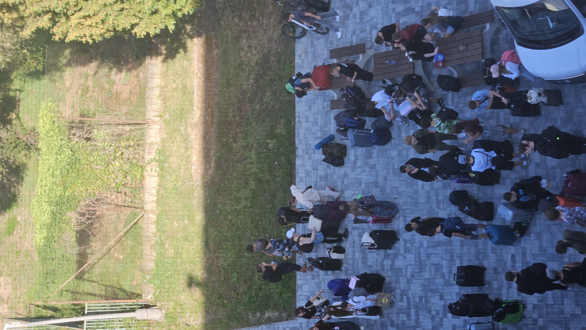 An aerial, high-angle shot captures a group of people gathered on a paved area, possibly a parking lot or courtyard, with luggage strewn around them. The ground is paved with rectangular gray tiles. In the background, there is a grassy area with some trees and a wire fence. The scene appears to be outdoors during the daytime, with shadows indicating sunlight.
The people, a mix of adults and teenagers, are dressed in casual clothing and are in various states of activity. Some are standing and talking, others are sitting on benches or the ground, and some are interacting with their luggage. The luggage includes various suitcases, backpacks, and duffel bags, predominantly in dark colors like black and gray, but also some blue, red, and green.
One person is loading luggage into the back of a white car visible in the lower right corner of the frame. Another person is sitting on a bicycle with a backpack. Several individuals are standing near picnic tables, some of which have bottles and containers on them.
The image does not provide specific visual cues to definitively identify the location as Prešov, Slovakia. However, the presence of picnic tables and what appears to be a public outdoor space with a car suggests a possible transportation hub, a park, or an area adjacent to a facility where groups might gather for travel or events. The overall atmosphere suggests a transient moment, perhaps people waiting for transportation or concluding a group activity.