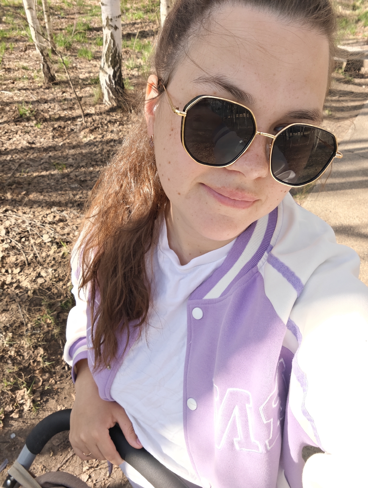 A woman is taking a selfie outdoors. She is wearing sunglasses, a white t-shirt, and a white and lilac jacket. She has her hair pulled back and is holding onto the handle of a stroller with her left hand, on which she is wearing a ring. The background includes trees and fallen leaves, suggesting a park or wooded area. The location is Chaikovsky, Russia.