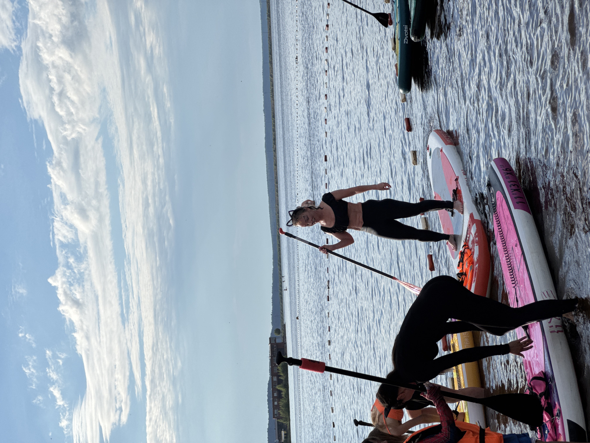 In this vertical outdoor shot, several people are engaged in paddleboarding on a calm body of water, likely a lake or river. The water is textured with small ripples, reflecting the sky. The sky itself is a pale blue, streaked with wispy white clouds, suggesting a bright, possibly breezy day. In the background, a distant shoreline features trees and buildings, including what appears to be an industrial complex with multiple windows and a tall chimney.
The primary subjects are the paddleboarders. One woman, wearing a black crop top and leggings and a black cat-ear headband, is standing on a pink and white paddleboard, holding a paddle. She is smiling and looking towards the camera. Another person in a black wetsuit is on a pink paddleboard, bending over and reaching for the water with one hand. Further in the foreground, partially visible, are other individuals also in wetsuits and life vests. The paddleboards are various colors, including pink, white, and orange.
The scene appears to be a recreational activity, possibly a lesson or group outing, given the number of people and paddleboards. The presence of buildings on the distant shore suggests a settled area, and the mention of Chaikovsky, Russia, places the activity within that geographical context. The lighting indicates it is daytime, likely afternoon given the quality of light. The expressions and activity suggest a fun, active pastime. Visible text on one paddleboard reads "76*15" and "DVOTSI".