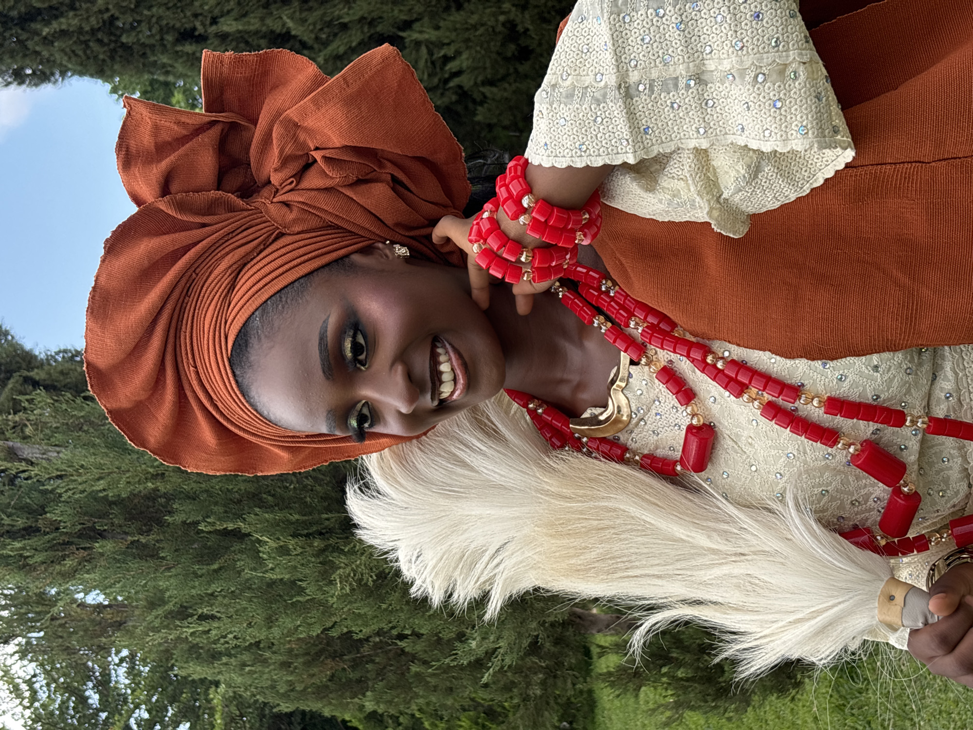The image shows a medium shot of a young Black woman in traditional attire, outdoors. She is wearing a rust-colored headwrap with a large, voluminous bow. Her outfit includes a cream-colored blouse with intricate white lace overlay adorned with tiny shimmering embellishments, and a rust-colored sleeveless vest. She has on multiple strands of bright red beaded necklaces and matching red beaded bracelets. The woman is holding a white feathery fan in her left hand. She is looking to her left with a wide, toothy smile and her makeup features prominent green eyeshadow. The background consists of lush green trees and a hint of blue sky. The lighting suggests it is daytime. The setting appears to be a garden or park area.