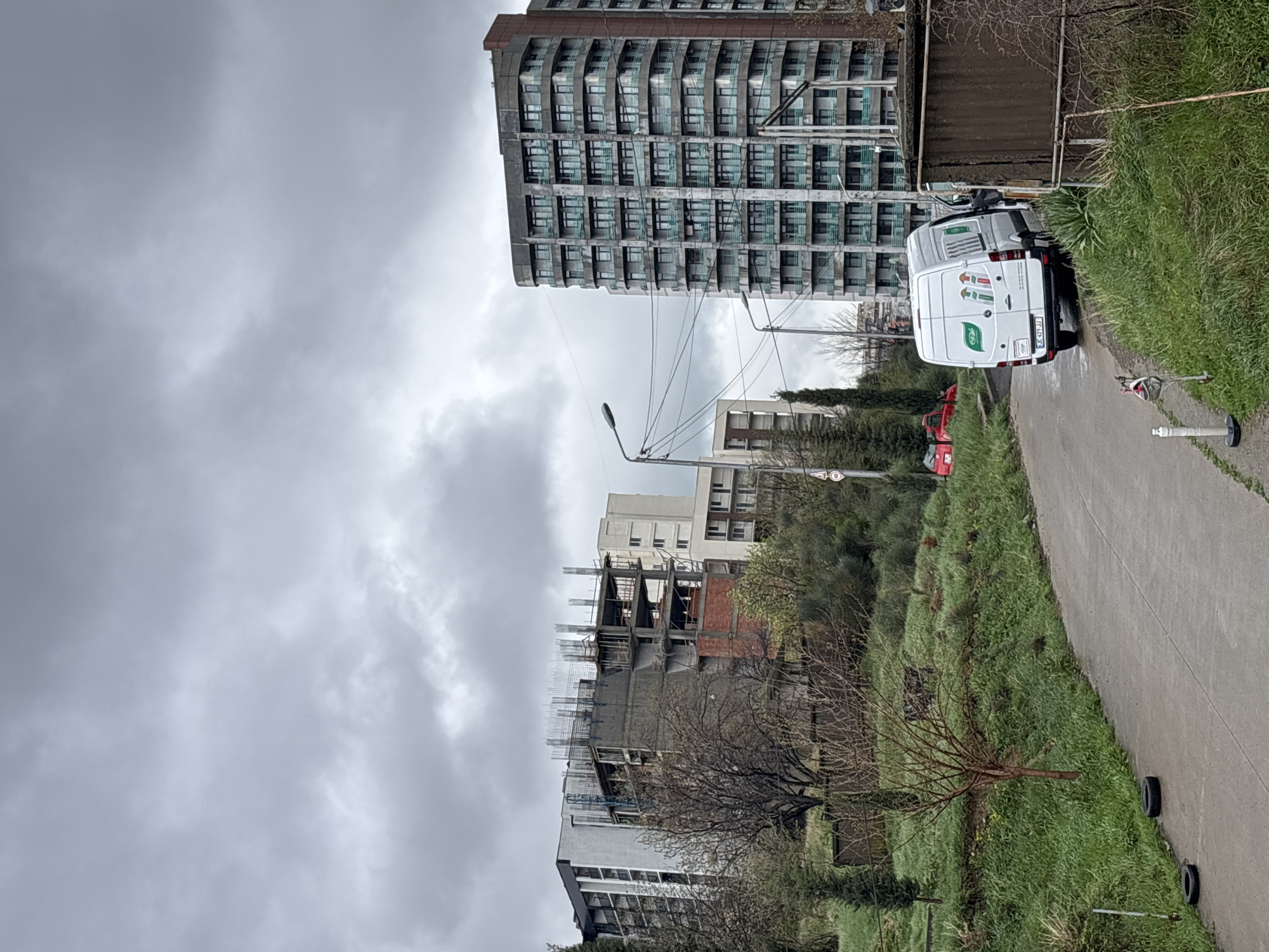 The image depicts a cloudy day in what appears to be a mixed urban and green space. In the background are several multi-story buildings, including an older, tall residential building with many balconies and a newer structure under construction. A white van is parked along a narrow asphalt road bordered by grassy areas. A red car is also partially visible behind the van. The environment includes overhead power lines and a few streetlights. The location is likely in Tbilisi, Georgia, based on the van's license plate and architectural styles of the buildings. The scene appears to be on the outskirts of the city center, with newer and older constructions juxtaposed.