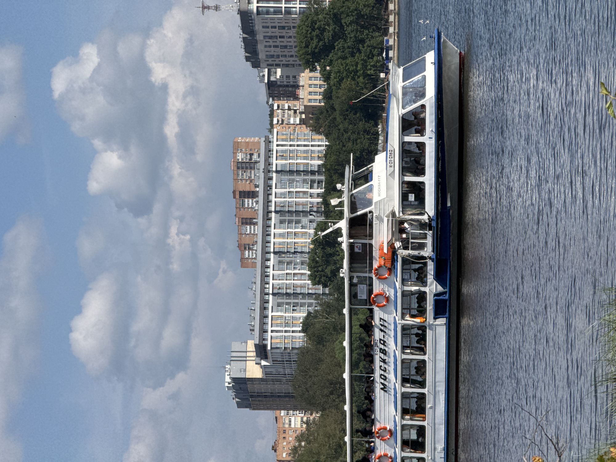 The image depicts a large white passenger boat, identified by the Cyrillic lettering "МОСКВА-117" on its side, sailing on a body of water. The boat is filled with passengers visible through its numerous windows. Orange life preservers are attached to the exterior of the boat. In the background, a dense line of green trees is visible, behind which are several modern multi-story buildings with a variety of architectural styles. The sky above is partly cloudy with large, fluffy white clouds against a blue background. The scene appears to be set on a river or lake within an urban environment, likely in Izhevsk, Russia, given the boat's designation. The water's surface is rippled, suggesting a slight breeze. The overall impression is of a sunny day with moderate cloud cover.