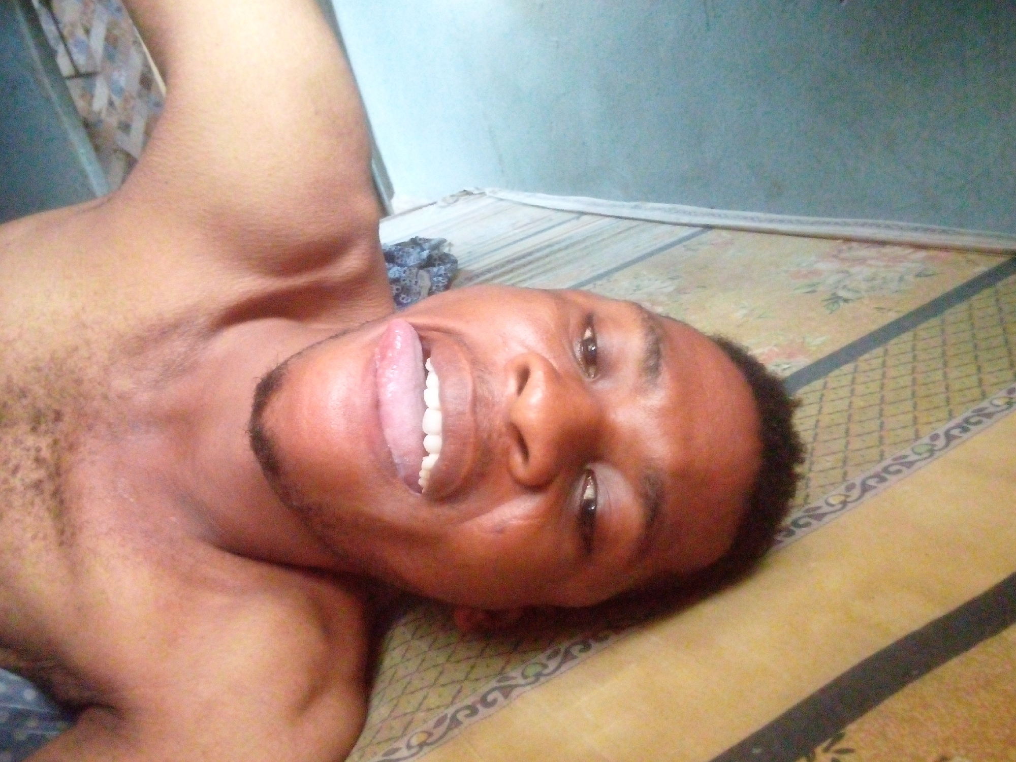 A close-up, angled shot features a shirtless, brown-skinned man lying on a patterned surface. His head is slightly tilted to the side, and his mouth is open, exposing his teeth and tongue. He has short, dark hair and a trimmed beard. The background includes a wall, visible in the upper left corner of the frame. The location is Unknown, Unknown.