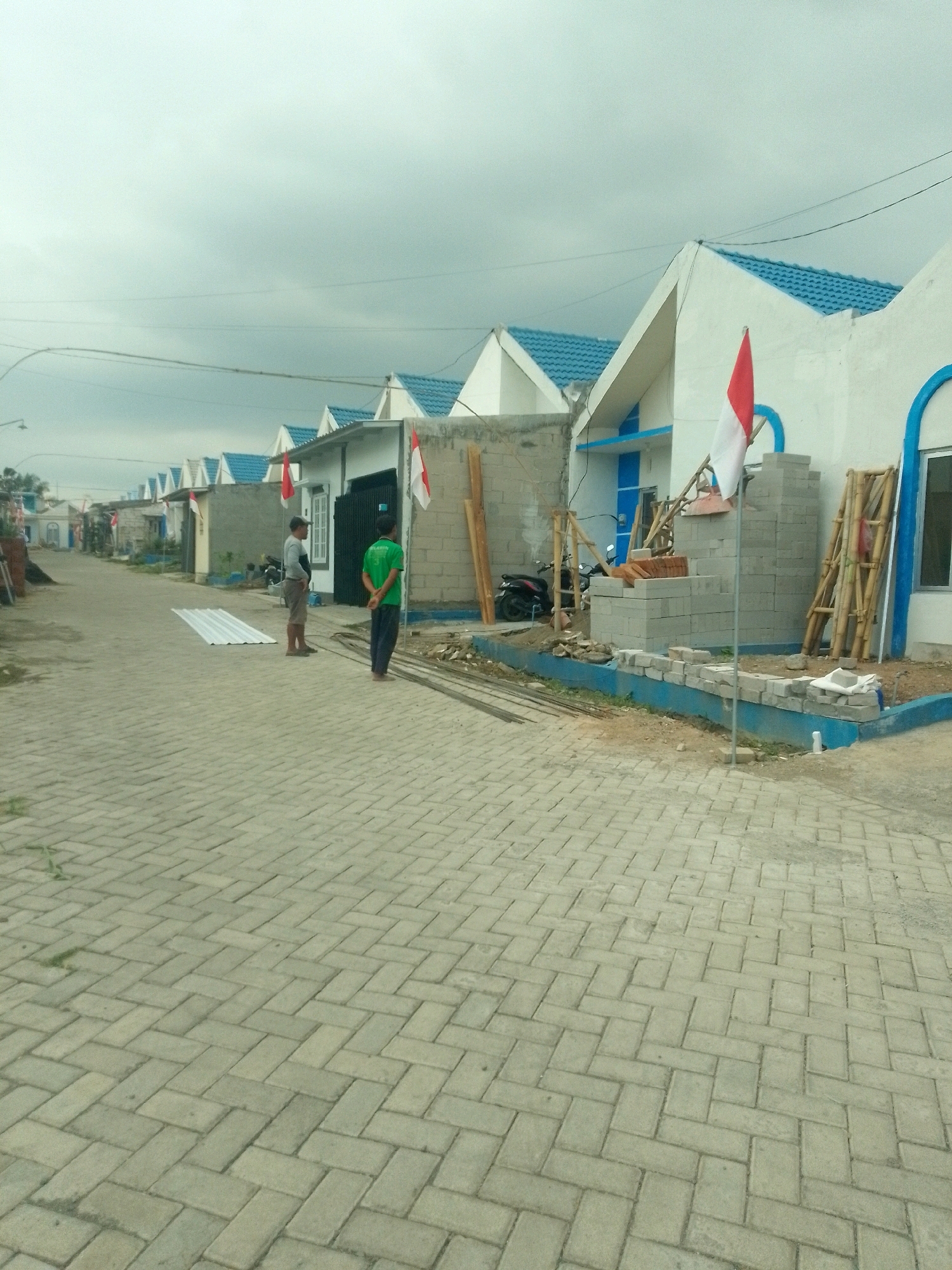 The image depicts a street scene in a residential area, likely under development, in Jember, Indonesia. The foreground is dominated by a paved road laid with interlocking bricks arranged in a herringbone pattern. To the left and extending into the distance, a row of houses with white walls and bright blue roofs is visible. Many of these houses appear to be under construction or recently completed, with building materials such as concrete blocks, lumber, and corrugated metal sheets present. Two men are standing in the street. One, wearing a grey shirt and shorts, stands near a stack of white corrugated metal. The other, in a green shirt and dark pants, is further down the street, facing away from the camera. Several Indonesian flags, red and white, are displayed on poles in front of some of the houses, suggesting a national holiday or patriotic observance. In the background, more houses and a few trees are scattered along the street, under an overcast sky. The overall impression is one of a developing residential neighborhood in Jember.
