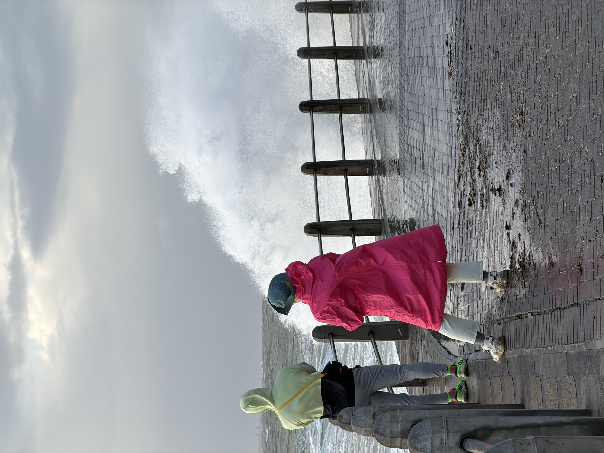 Two people wearing rain gear stand behind a metal railing at the Sea Point Promenade in Cape Town, South Africa. A wave is crashing near the railing. The person closest to the camera wears a lime green hooded raincoat, gray pants, and neon green and black athletic shoes. The other person wears a pink raincoat, light-colored pants, and white and tan athletic shoes. The pavement is wet.