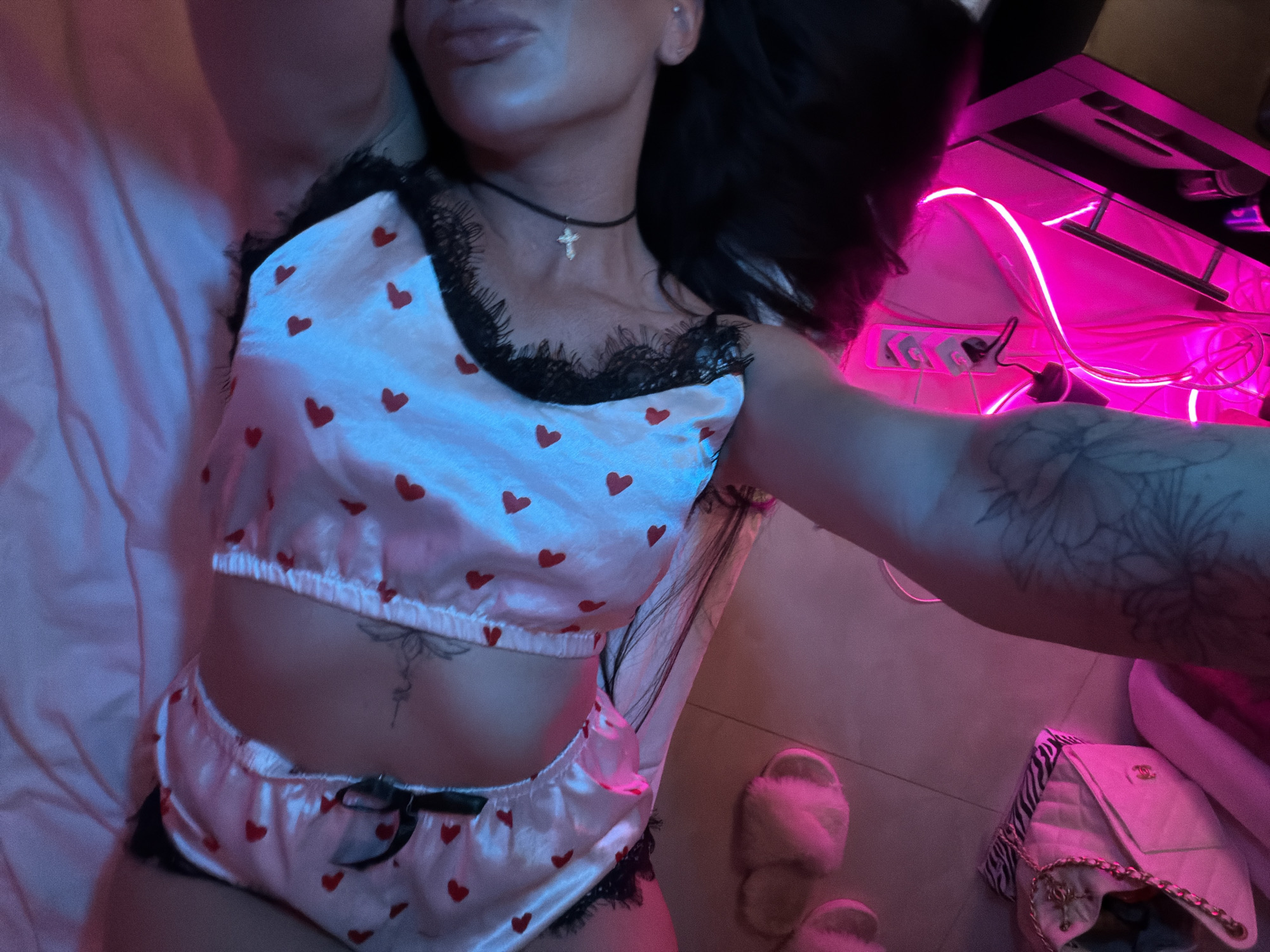 An overhead shot captures a person, likely a woman, lying down indoors, seemingly taking a selfie with her tattooed right arm outstretched. The scene is set in what appears to be a private room in Boreti, Montenegro, illuminated by a prominent neon pink LED strip light on the right, casting a vibrant glow that indicates an evening or nighttime setting.
The subject's face is partially visible, showing dark hair and full lips. She wears a two-piece satin pajama set adorned with a pattern of red hearts on a white background, accented with black lace trim along the edges of the crop top and short bottoms. A black choker with a small, silver cross pendant is worn around her neck. Distinctive tattoos include a floral or foliage design on her right arm and a delicate, intricate symbol positioned centrally on her abdomen, just below the crop top.
She lies on what appears to be a light-colored bed or mattress, with the bedding visible on the left. To the right, on a light-toned tiled floor, a pair of fluffy, light-colored slippers are present, alongside a white quilted handbag with a gold chain strap and a classic designer-style clasp, reminiscent of a Chanel bag. In the upper right background, a dark desk or shelf unit is visible, beneath which several power adapters and cables are plugged into a power strip, all bathed in the strong pink illumination. The image conveys a relaxed and personal atmosphere.