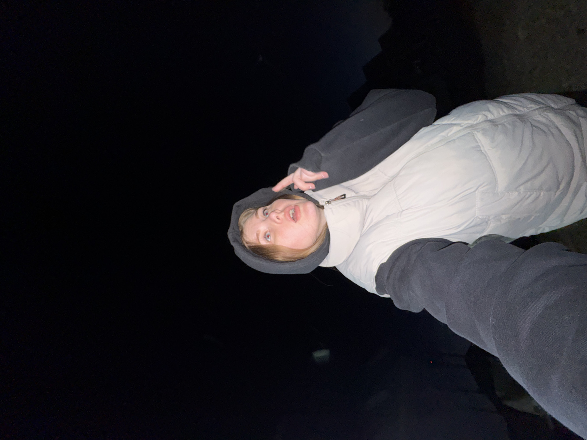 A person is posing for a selfie at night. The person is wearing a hooded sweatshirt and a white puffy vest. They are holding up two fingers. The location appears to be outdoors, with a dark background. The location, Gornyi (Dvurechenskoe S/P), Russia, cannot be verified from the image.
